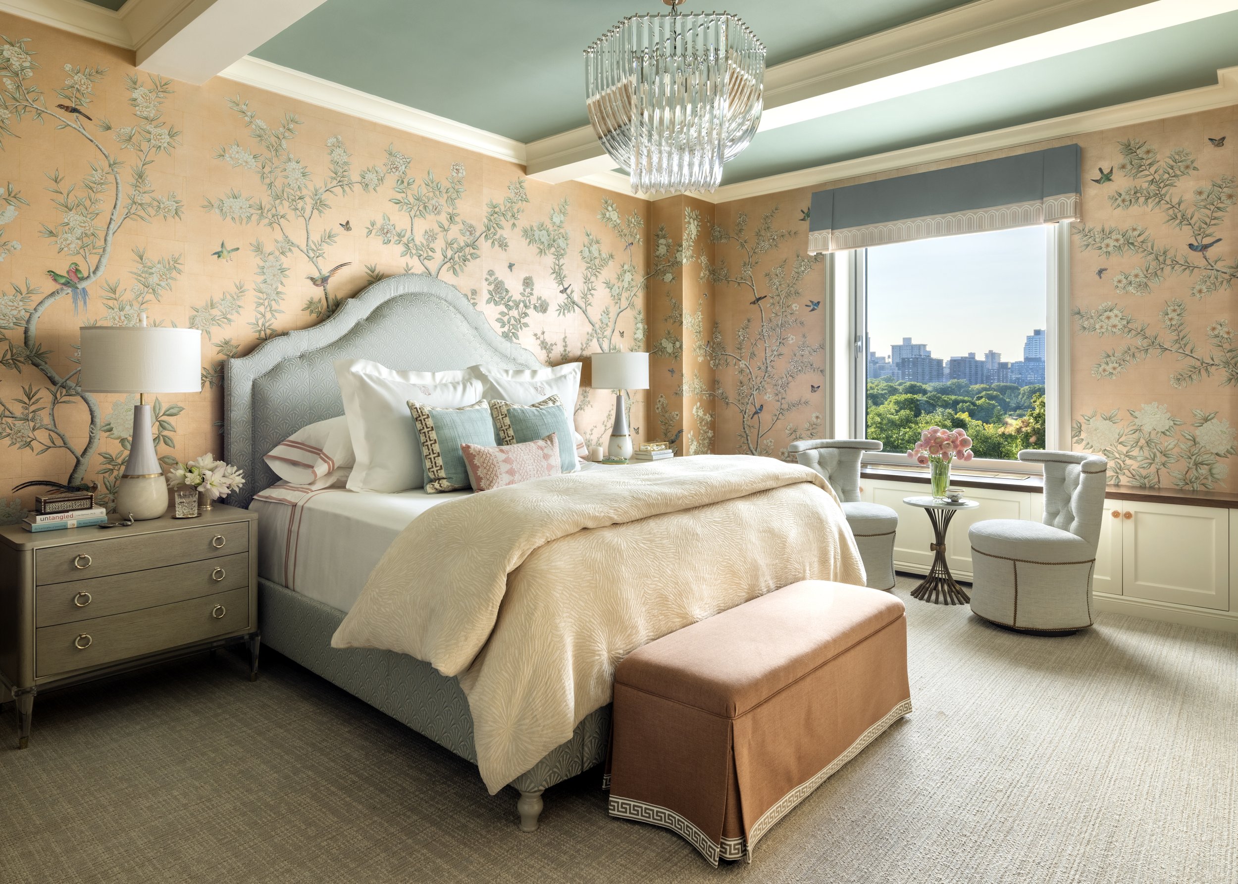 A bedroom with pastel green ceiling, peach floral wallpaper with birds and butterflies, a bed with a scalloped upholstered headboard, multiple pillows, bedside lamps, a small round table with a pink flower bouquet, and a large window showing a city skyline and green trees.