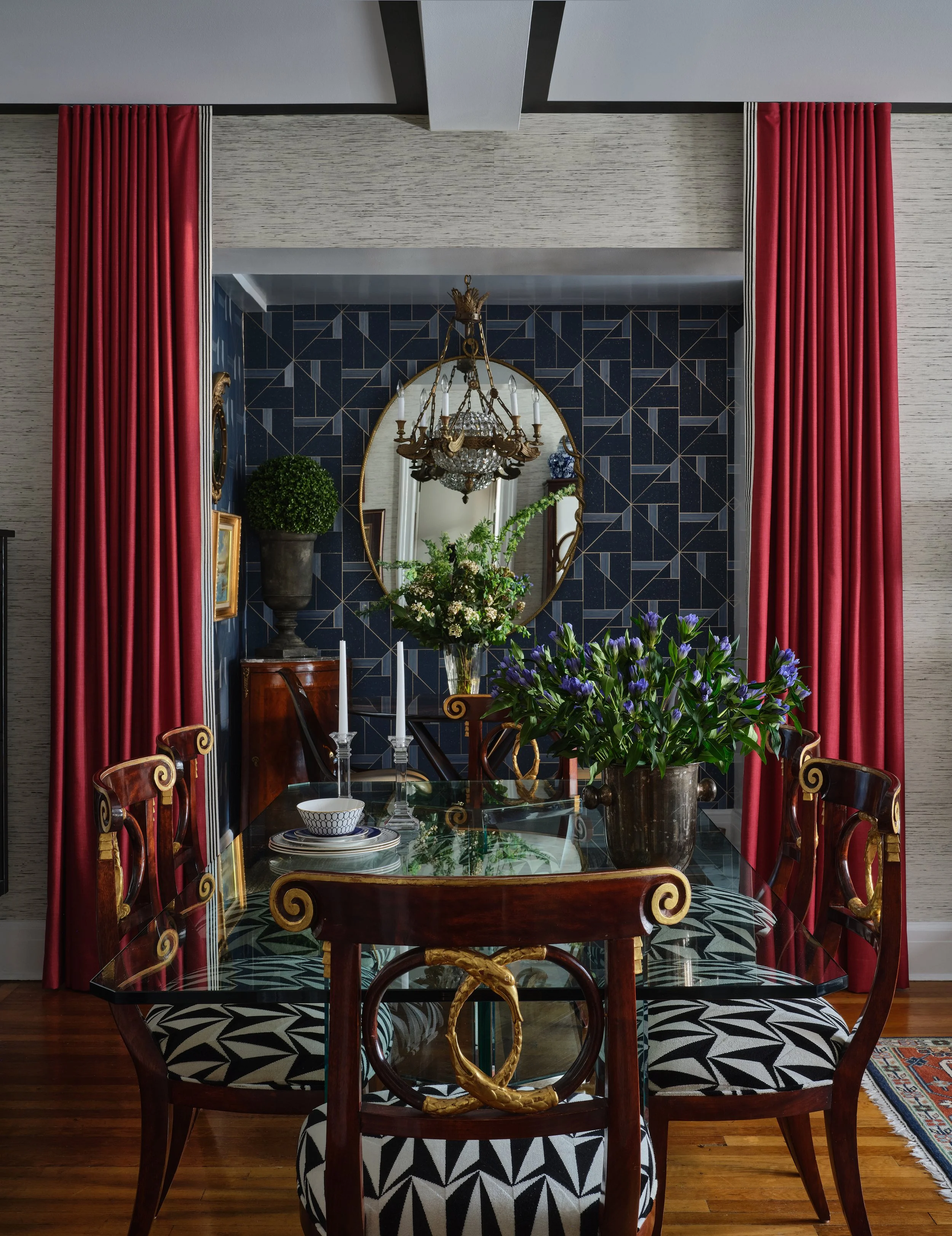 A dining room with a glass-topped table surrounded by wooden chairs with black and white patterned cushions, decorated with a large potted plant, a vase with purple flowers, and a candlestick holder. In the background, a mirror and chandelier are vis