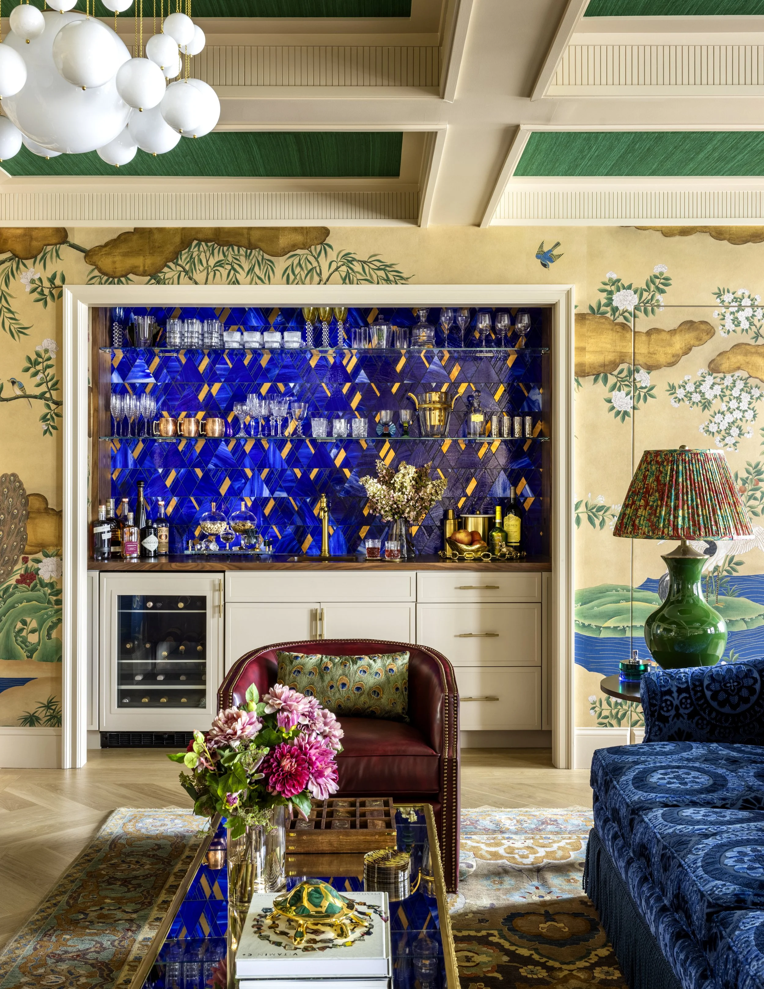 A luxurious interior with colorful and ornate decor, a floral arrangement on a glass-top coffee table, a burgundy armchair, a blue velvet sofa, a green vase, a tray with snacks, and a decorative lamp. There is a built-in bar with glassware and bottles, and a wall with a mural of flowers, clouds, and a bird.