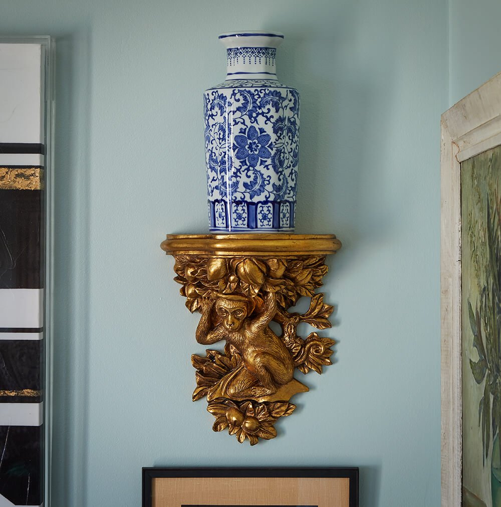A blue and white porcelain vase placed on a decorative golden wall mount shaped like a monkey holding onto leaves.