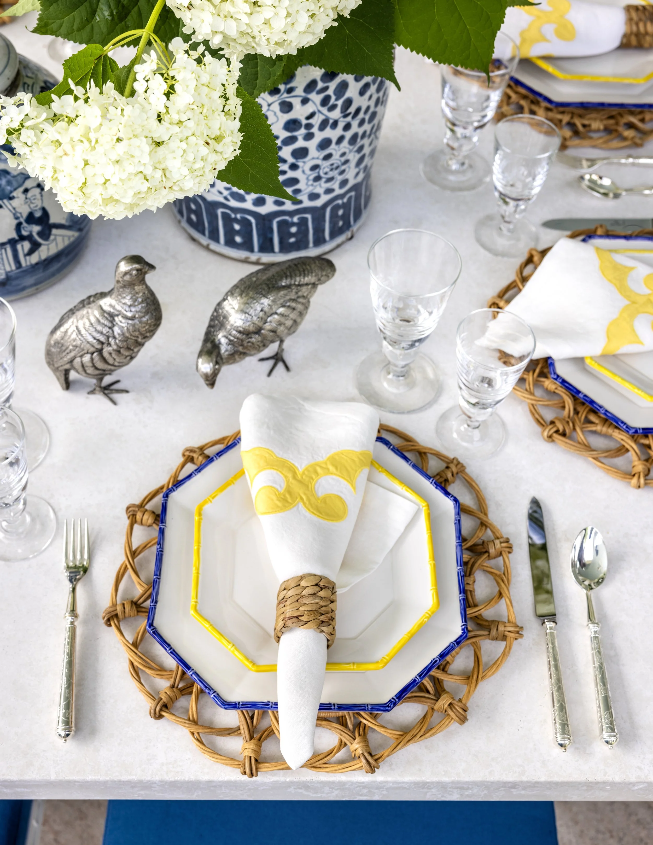Table setting with a woven basket-like charger plate, a white napkin with yellow and white napkin ring, silverware, clear glass goblets, a floral centerpiece with white hydrangeas in a blue and white patterned vase, and decorative metal bird figurine