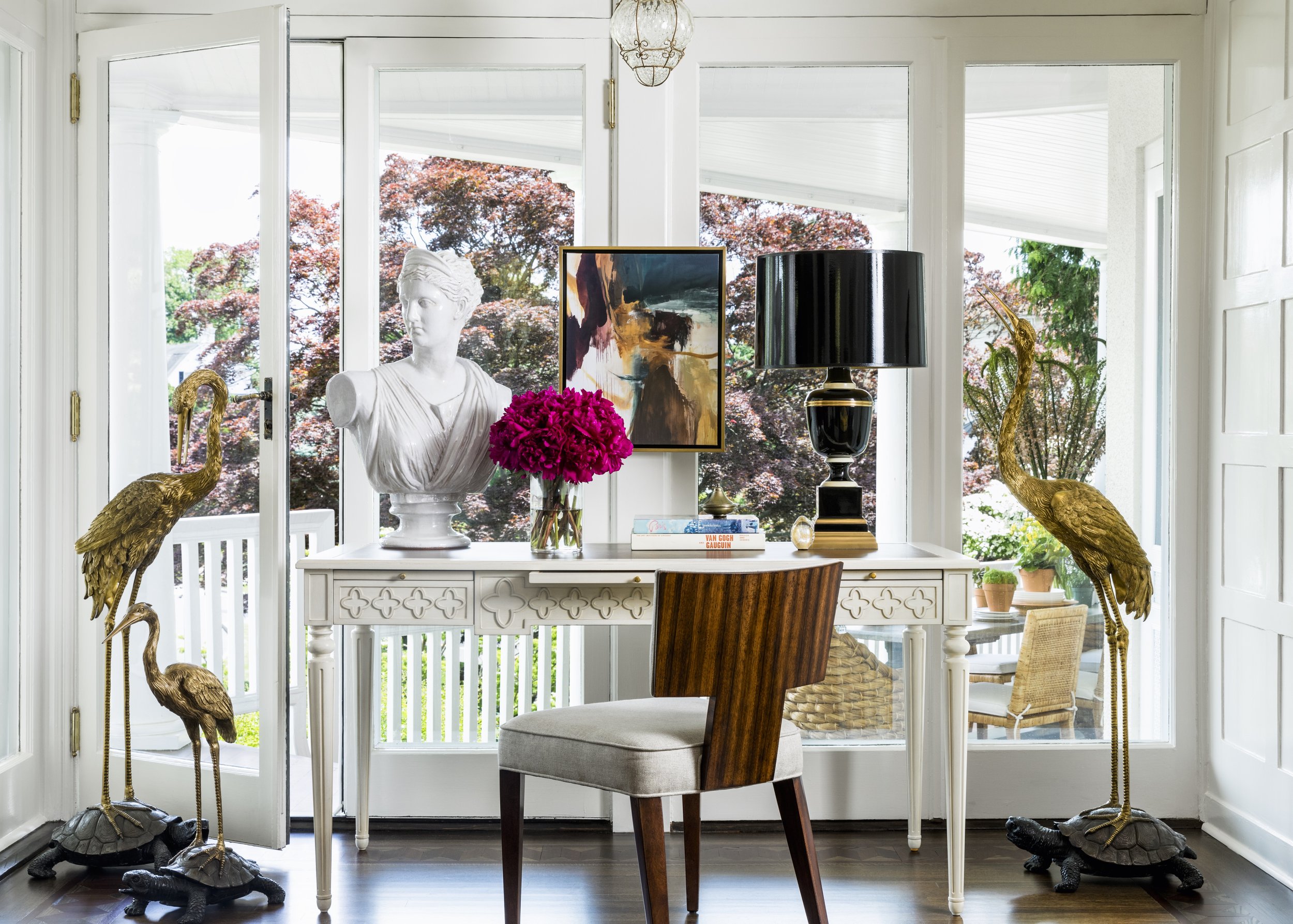An interior space with a white console table adorned with decorative items, including a white bust, pink flowers in a glass vase, a black and gold table lamp, and books. Flanking the table are two tall gold crane sculptures with turtle bases. Behind 