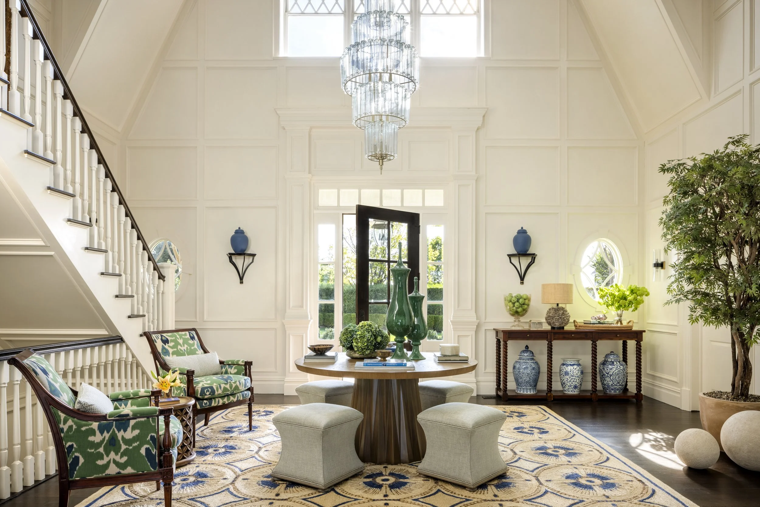 A spacious, elegant living room with white paneled walls, high ceilings, and large windows. A circular wood table with green vases, surrounded by chairs and ottomans. Decorative blue and white vases, a side table with a lamp, and a tall potted plant 