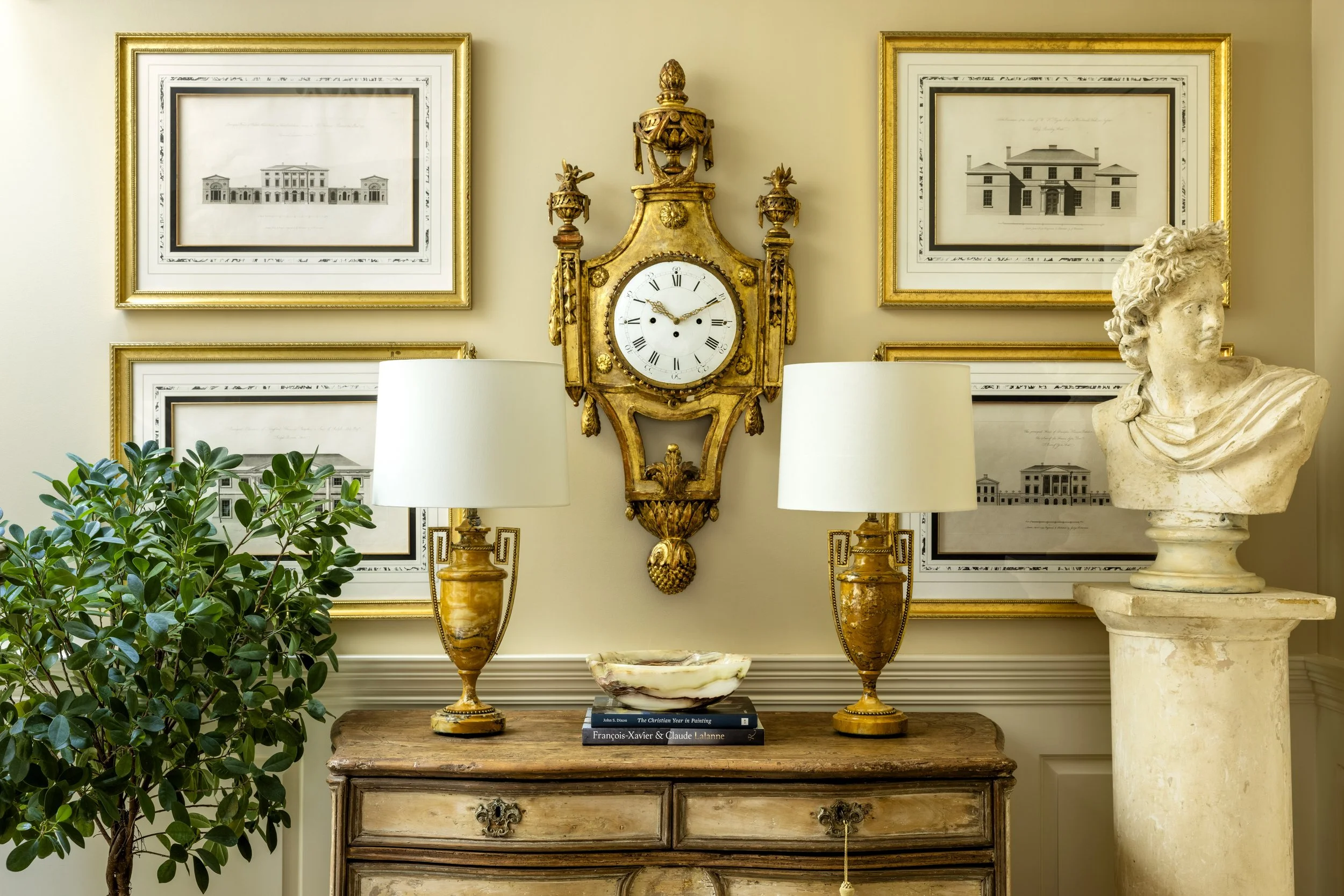 Vintage gold wall clock centered between two framed architectural blueprints, flanked by table lamps, with a bust sculpture on the right and green plants on the left, all on a wooden console table.