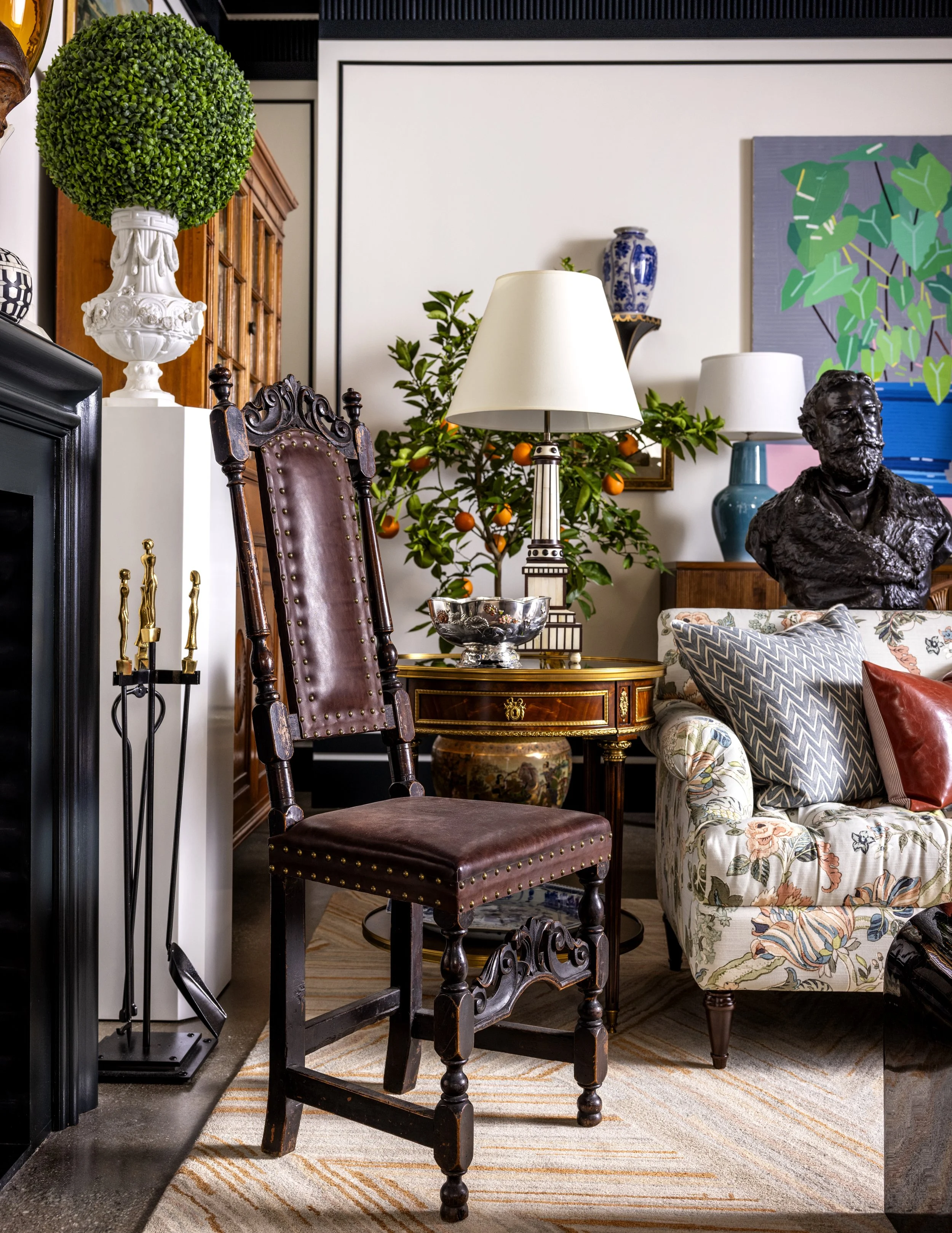 Interior of a living room with antique chairs, a table with a silver bowl, lamps, a bust sculpture, painting, and decorative plants.
