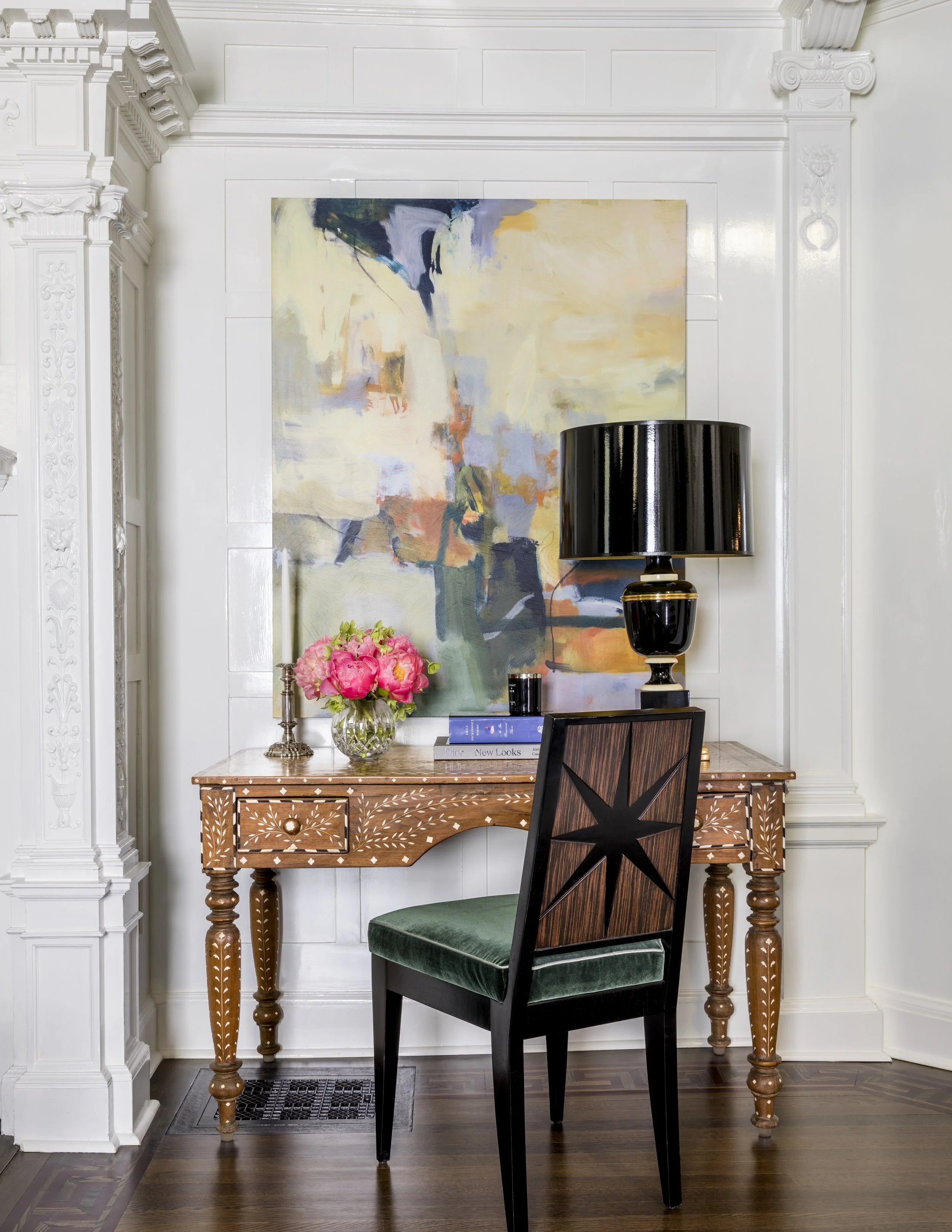 Interior room with ornate white woodwork, a wooden desk, a black and wood chair, a lamp, framed artwork, a vase of pink flowers, and books.