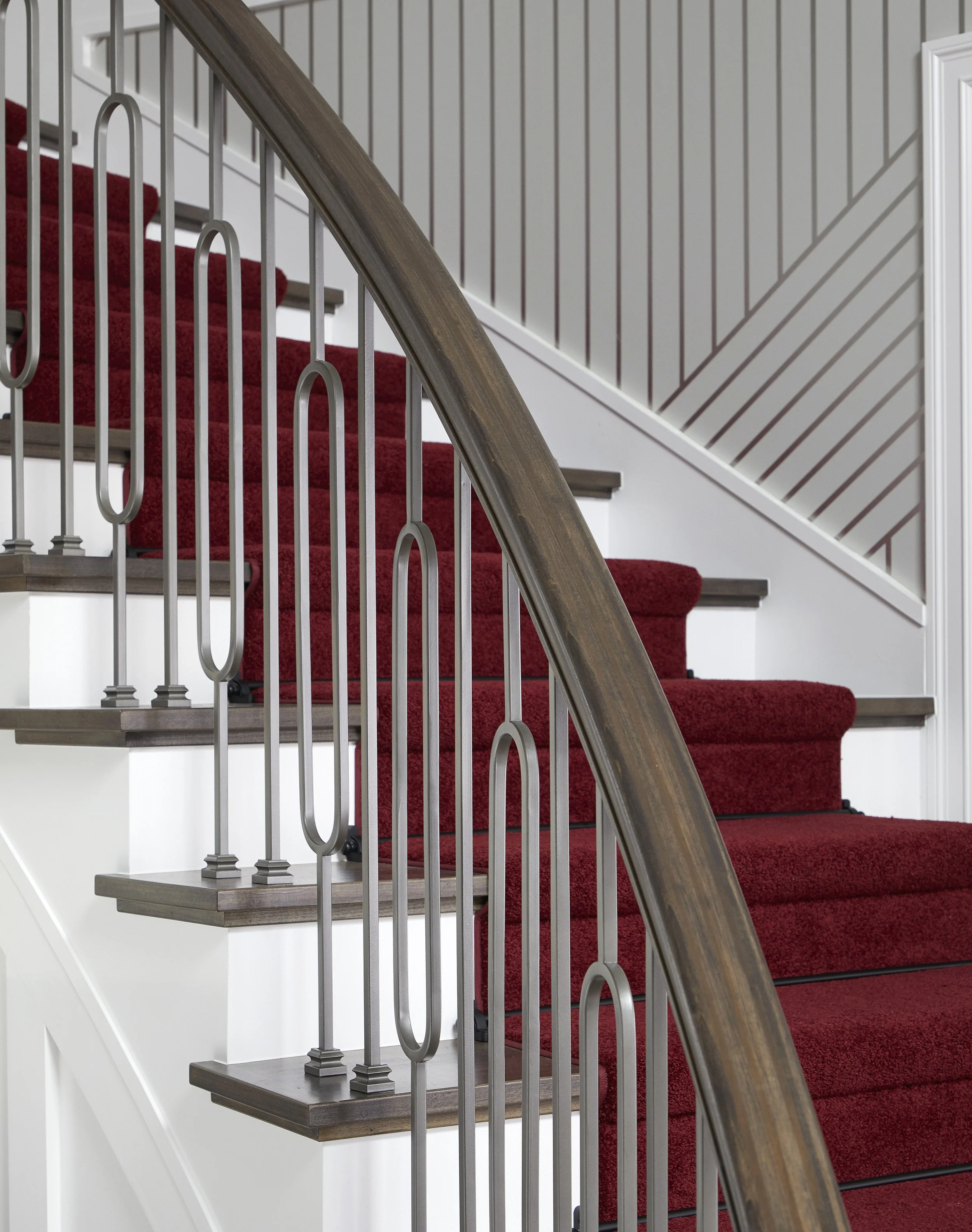 Interior view of a staircase with red carpeted steps, a wooden handrail, and decorative metal balusters, against a wall with wood paneling.