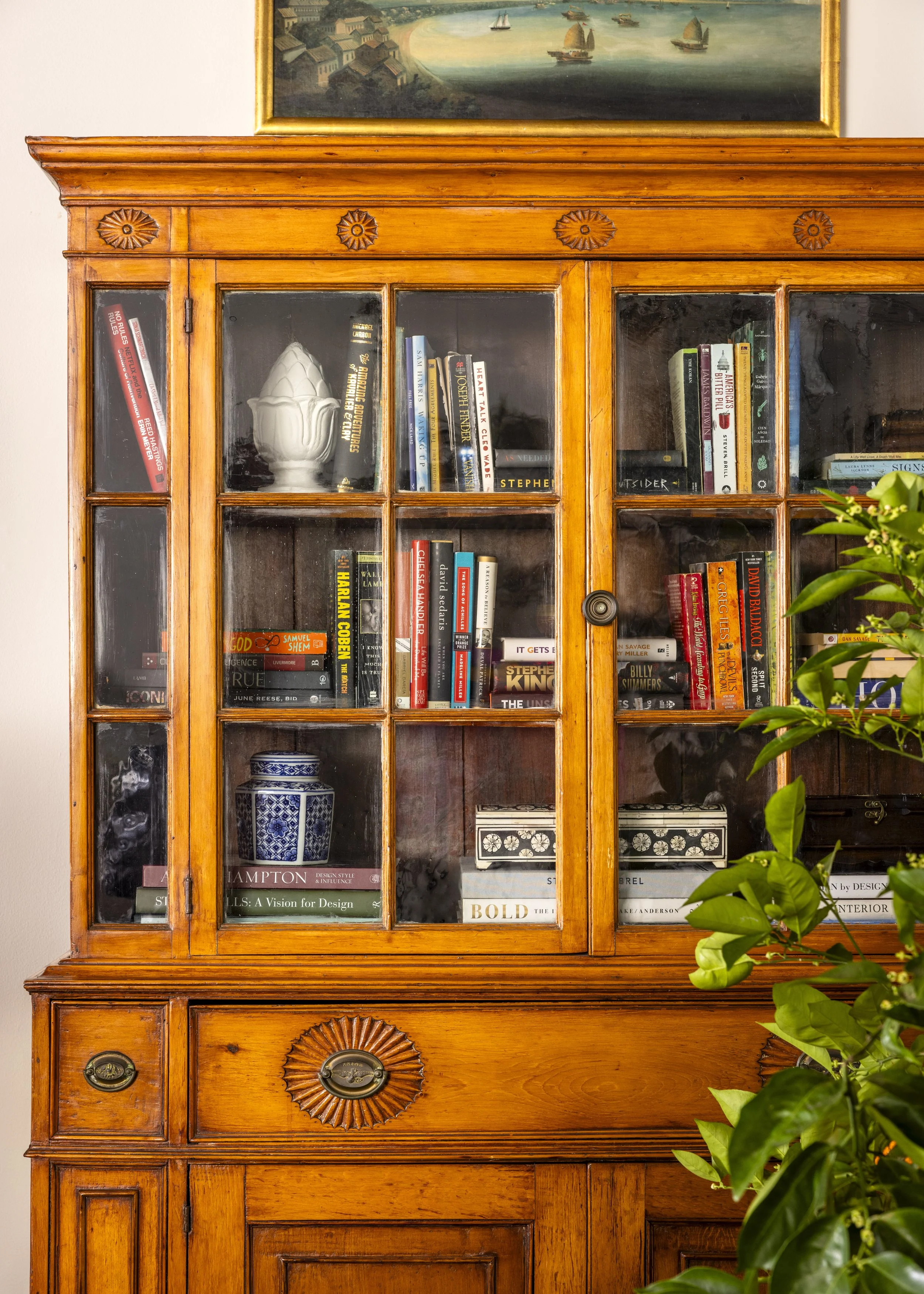 A wooden bookshelf with glass doors, filled with books, decorative items, and a ceramic artichoke on the middle shelf. There is a plant with green leaves partially visible on the right side. A landscape painting of boats on water hangs above the book