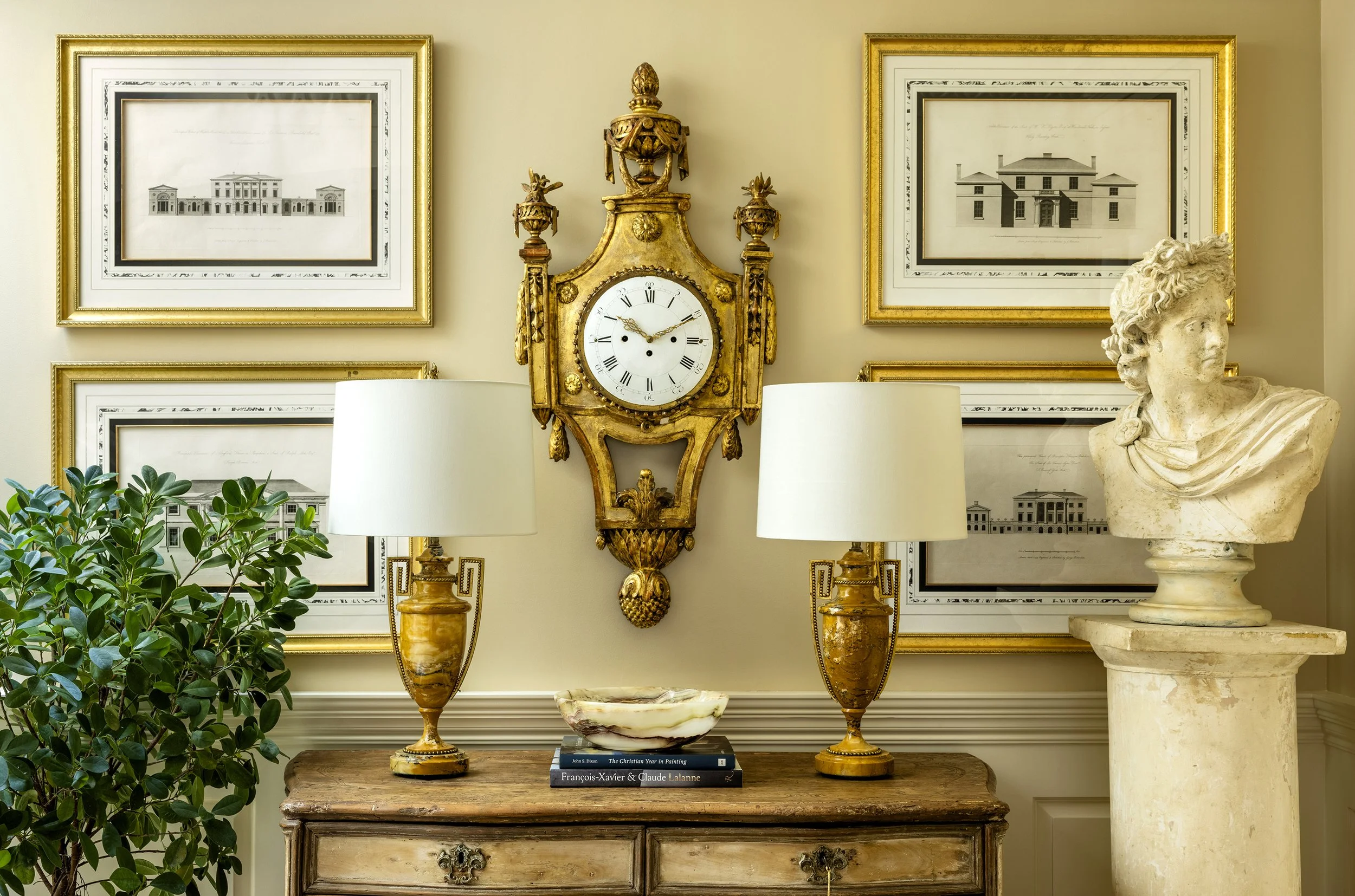 Elegant interior decor featuring a large antique wall clock with gold accents, framed architectural drawings, a classical bust sculpture, two table lamps, potted green plant, a wooden table, and a stack of books.
