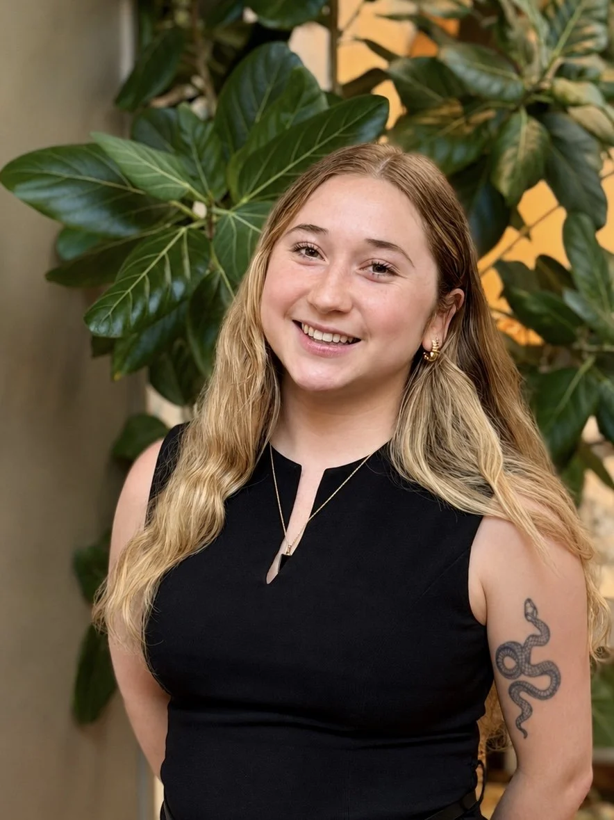 A smiling young woman with long, wavy blonde hair, wearing a black sleeveless top, gold earrings, and a necklace, standing in front of large green leafy plants.