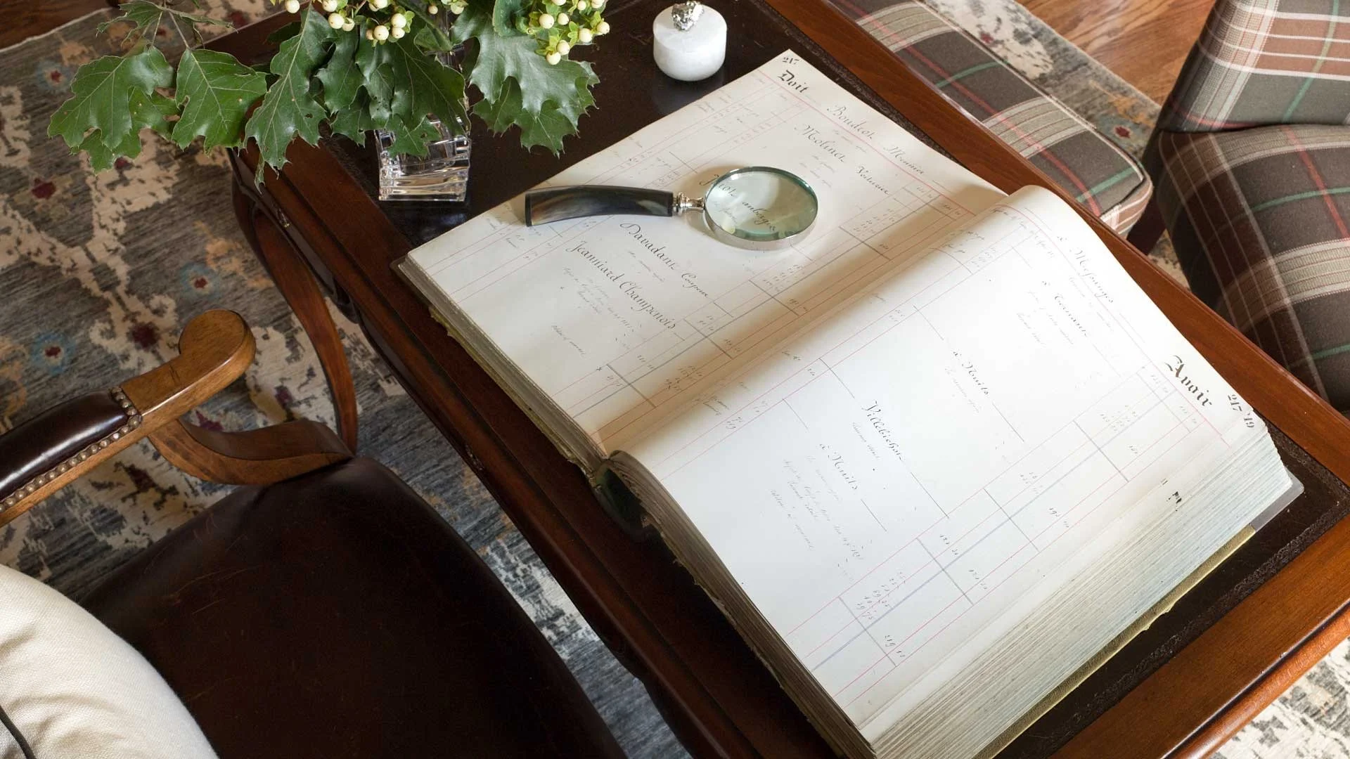 Open ledger book on a wooden table with a magnifying glass resting on it. There is a vase with green holly leaves and white berries on the table, along with a small white vase. A plaid armchair and a patterned rug are visible nearby.