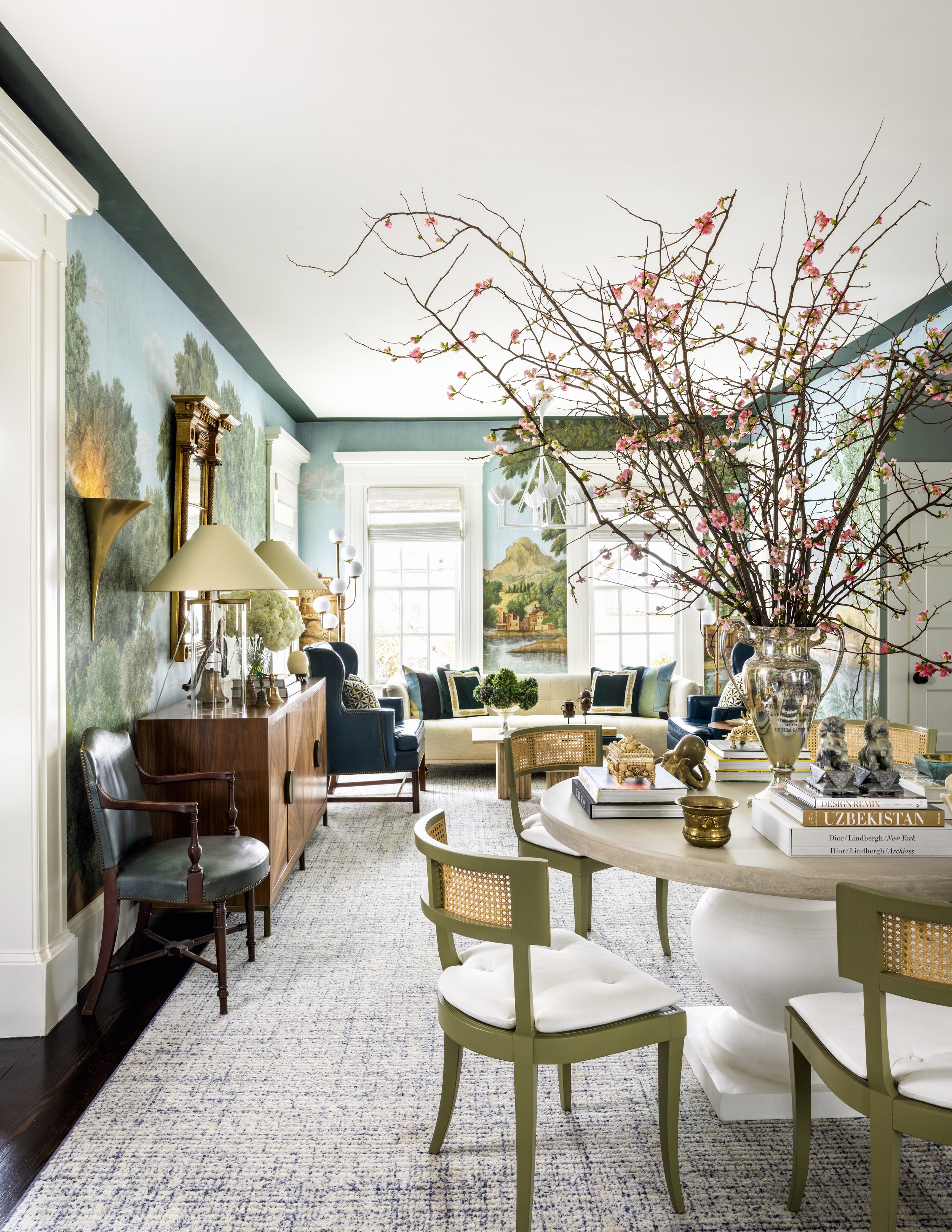 Living room with a large vase of cherry blossom branches on a table, blue chairs, painting of a landscape, and nautical-themed decor.