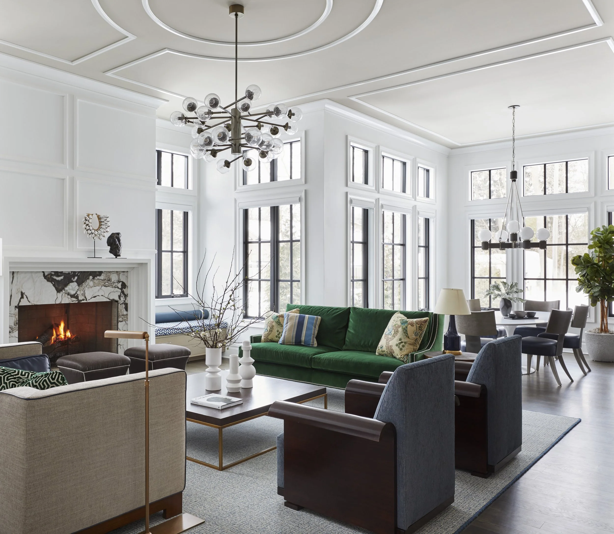 Modern living room with a white ceiling featuring crown molding, large windows, a fireplace, two chandeliers, and various seating including a green velvet sofa, armchairs, and ottomans.
