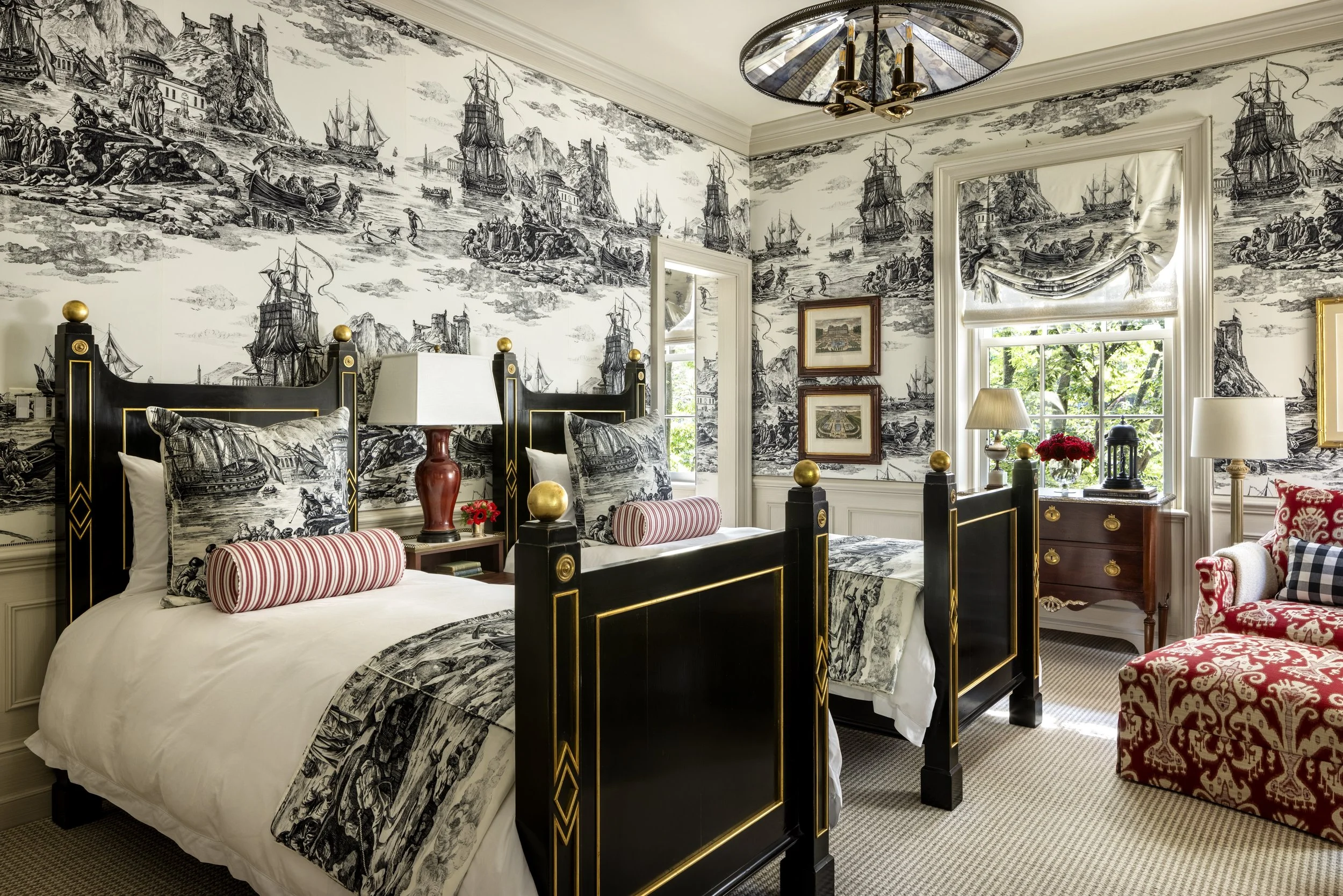 A bedroom with black and gold bed frames, detailed black and white nautical-themed wallpaper, a window with a valance, and various furniture and decor including lamps, artwork, and a red patterned armchair.
