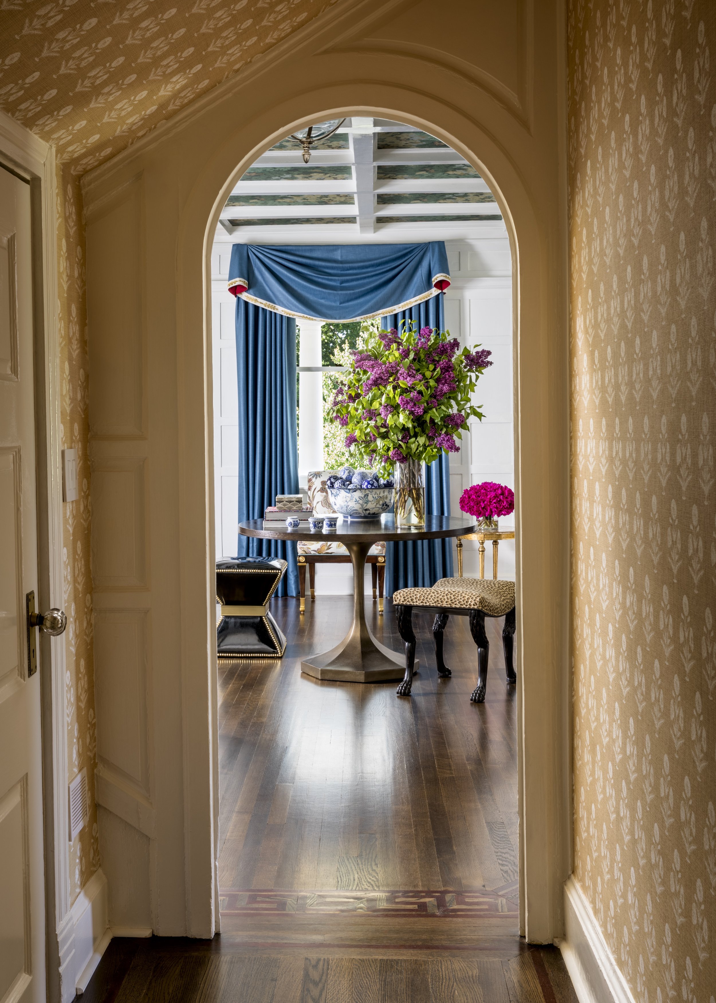 View through an arched doorway into a sunlit room with a round table, large floral arrangement, blue curtains, and patterned wallpaper.