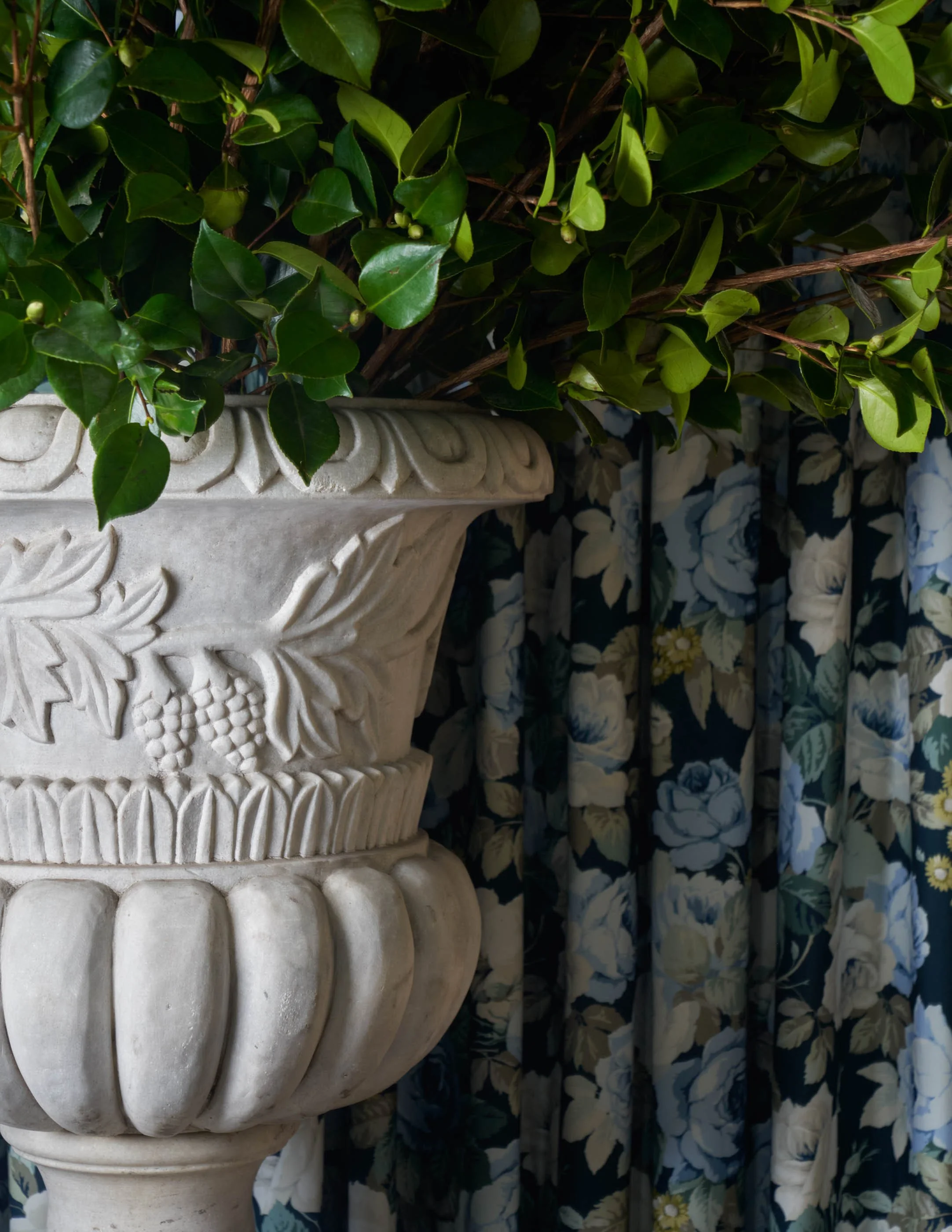 A decorative beige stone urn with intricate patterns, filled with green leafy plants, placed in front of a floral curtain with blue and white flowers.