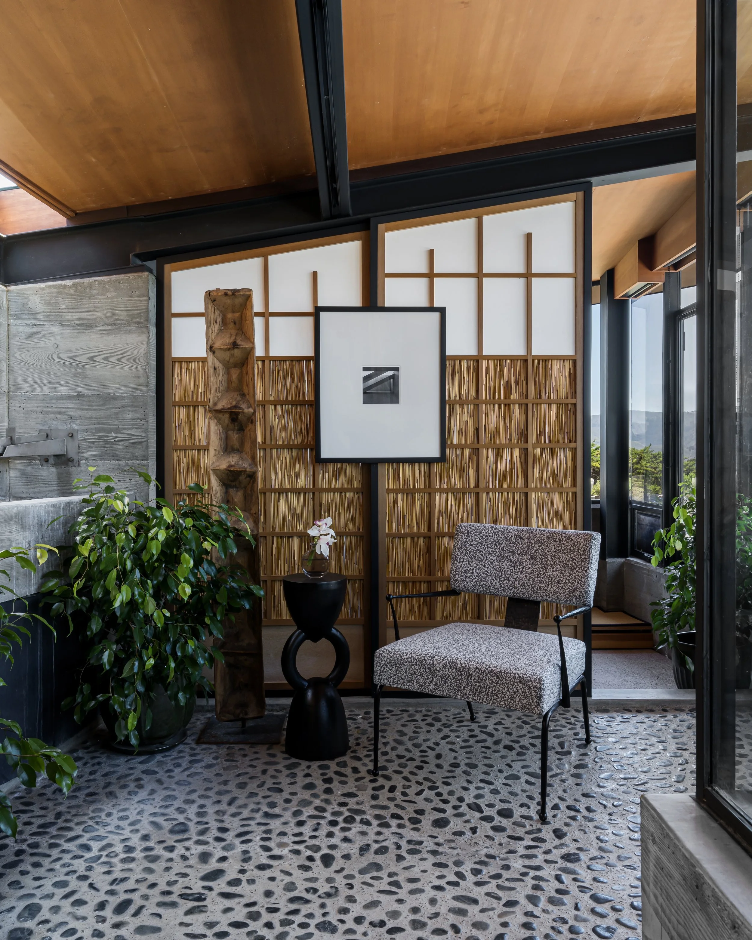 Interior of a room with a large plant on the left, a black sculpture and a small table in the center, and a patterned chair by the window on the right. The room has wooden and bamboo decor, with large windows letting in natural light, and a pebble st