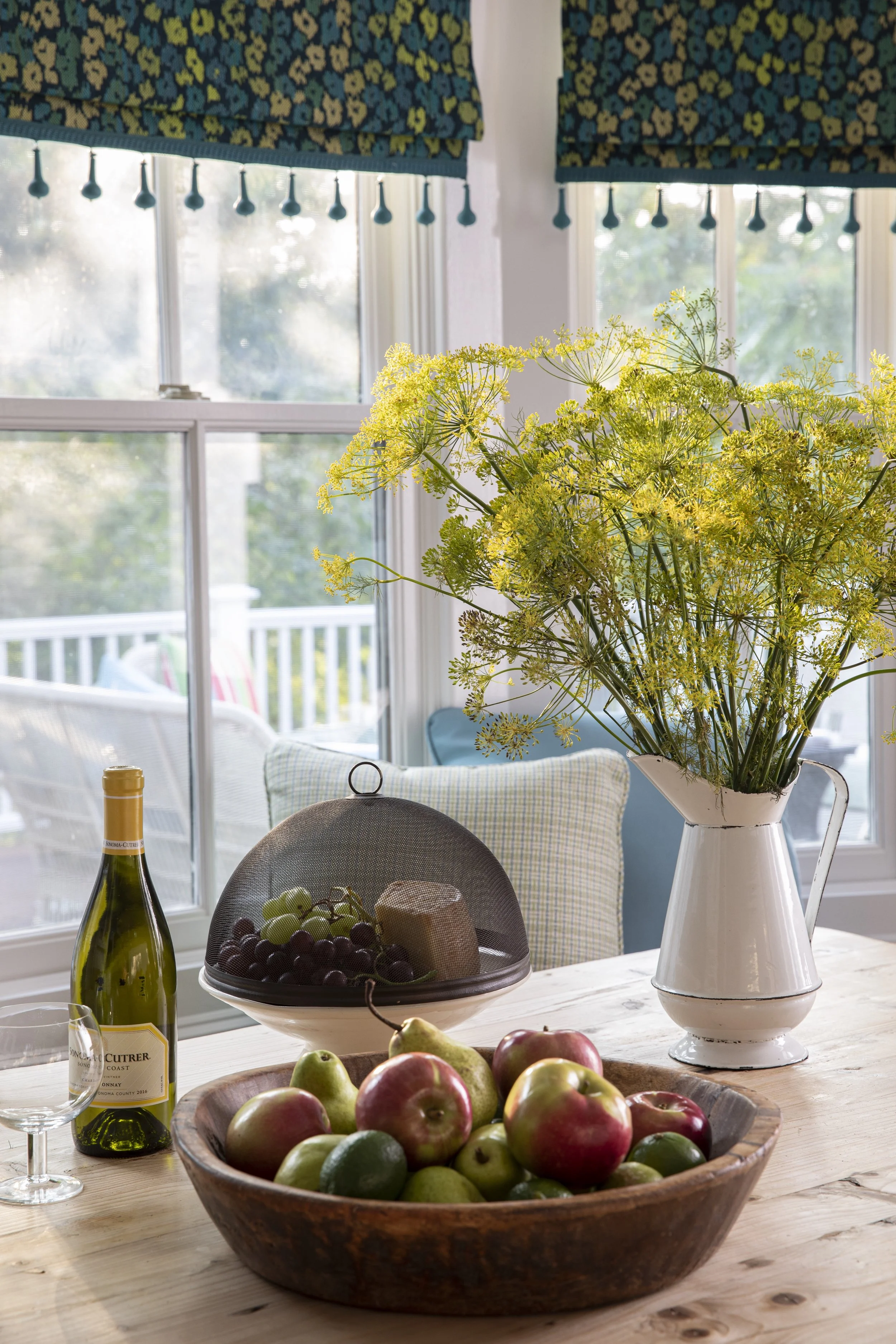 A wooden bowl filled with apples and limes on a table, with a bottle of wine, a glass, and a covered tray of grapes and cheese nearby. A large white vase with yellow flowers sits on the table. In the background, there are window curtains, a window wi