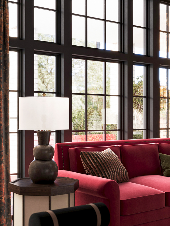 Interior of a room with large grid windows, a red sofa with patterned and green cushions, a table with a large lamp, and some plants outside visible through the windows.