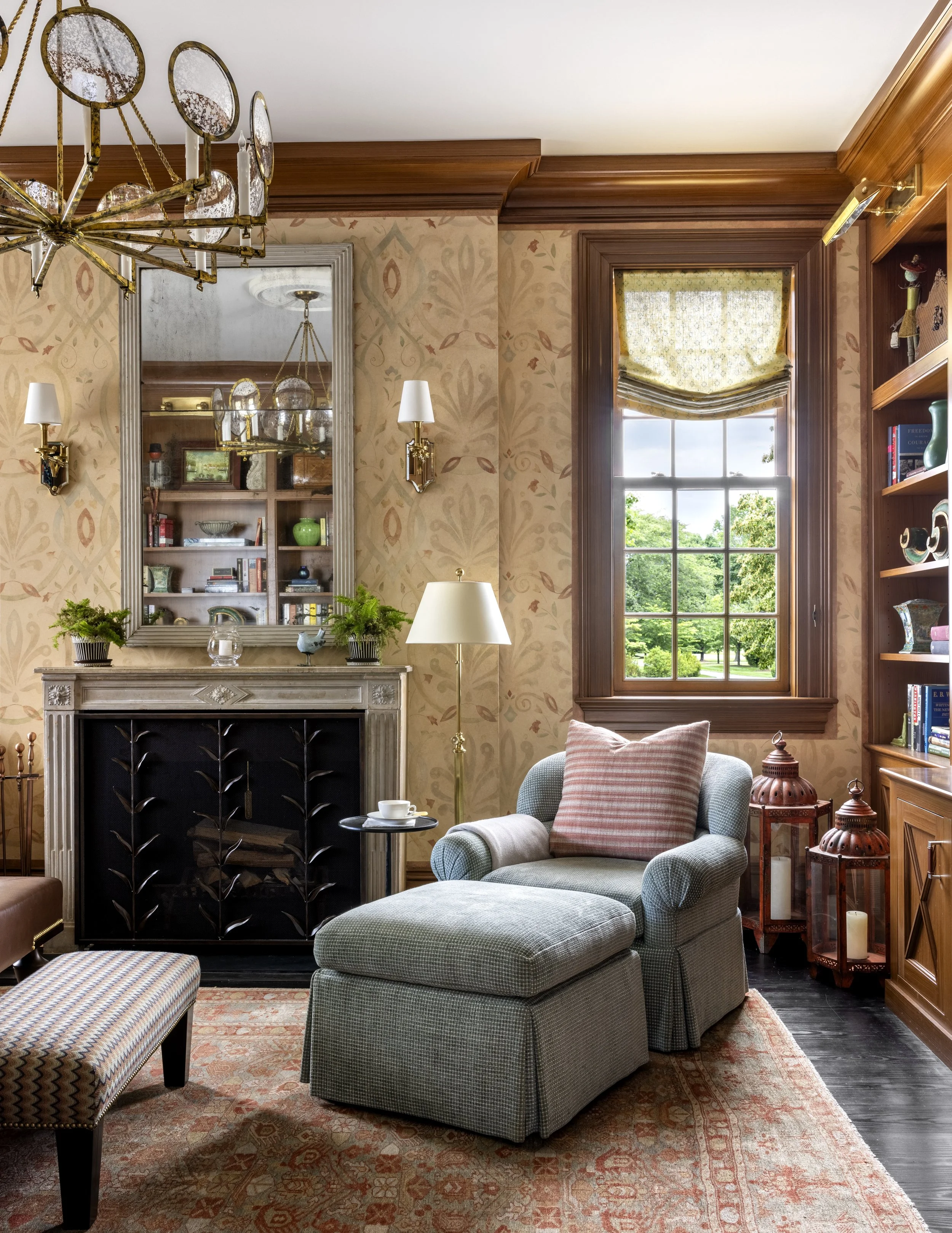 A cozy living room with a window showing greenery outside, a grey armchair with a pink pillow, a fireplace with a mirror above it, a floor lamp, and a bookshelf filled with books and decorative items, near a patterned area rug.