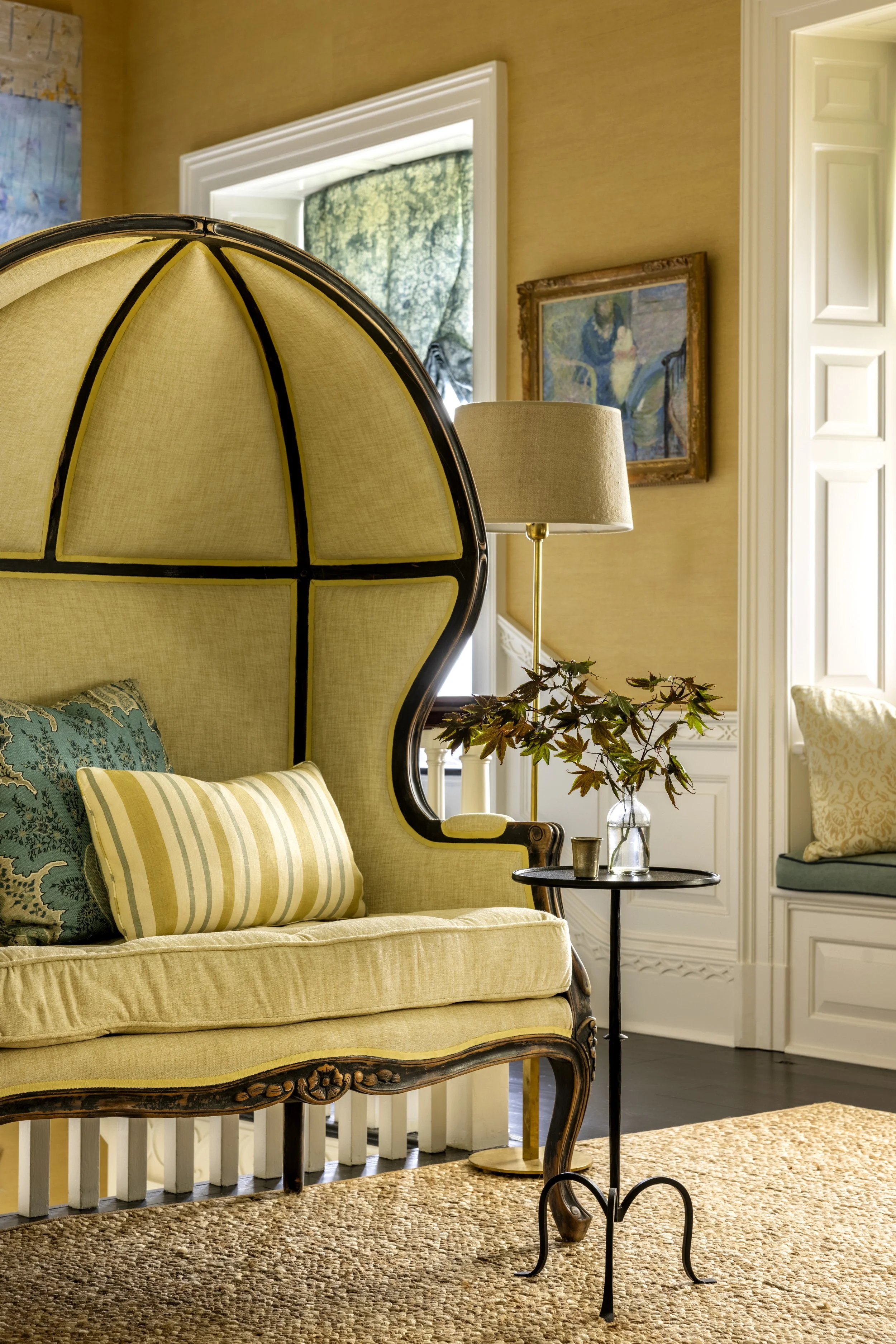 A cozy living room corner with a vintage yellow sofa, patterned cushions, a black side table with a glass vase and plants, a standing lamp, framed artwork, window with green curtains, and beige walls.