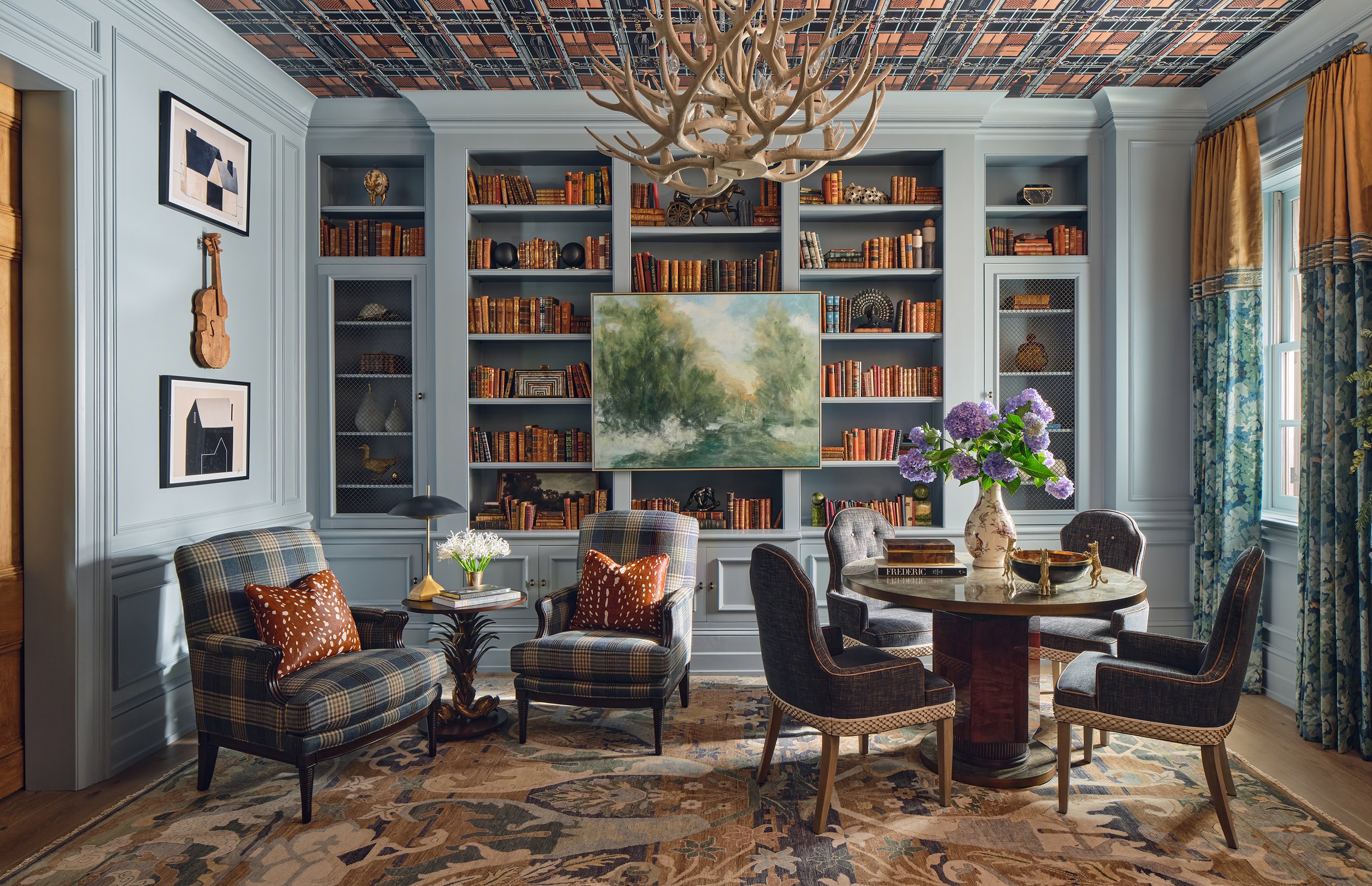 A cozy living room with light blue walls and a built-in bookshelf filled with books and decorative objects. There are several armchairs and a round wooden table with a vase of purple hydrangeas. The ceiling has a patterned design, and there's a flora