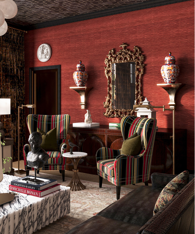 A stylish living room with two high-back, striped armchairs with green pillows, a marble-topped coffee table with books and a bust, a dark sofa with pillows, and a red textured wall decorated with a large ornate mirror and two decorative vases on ped