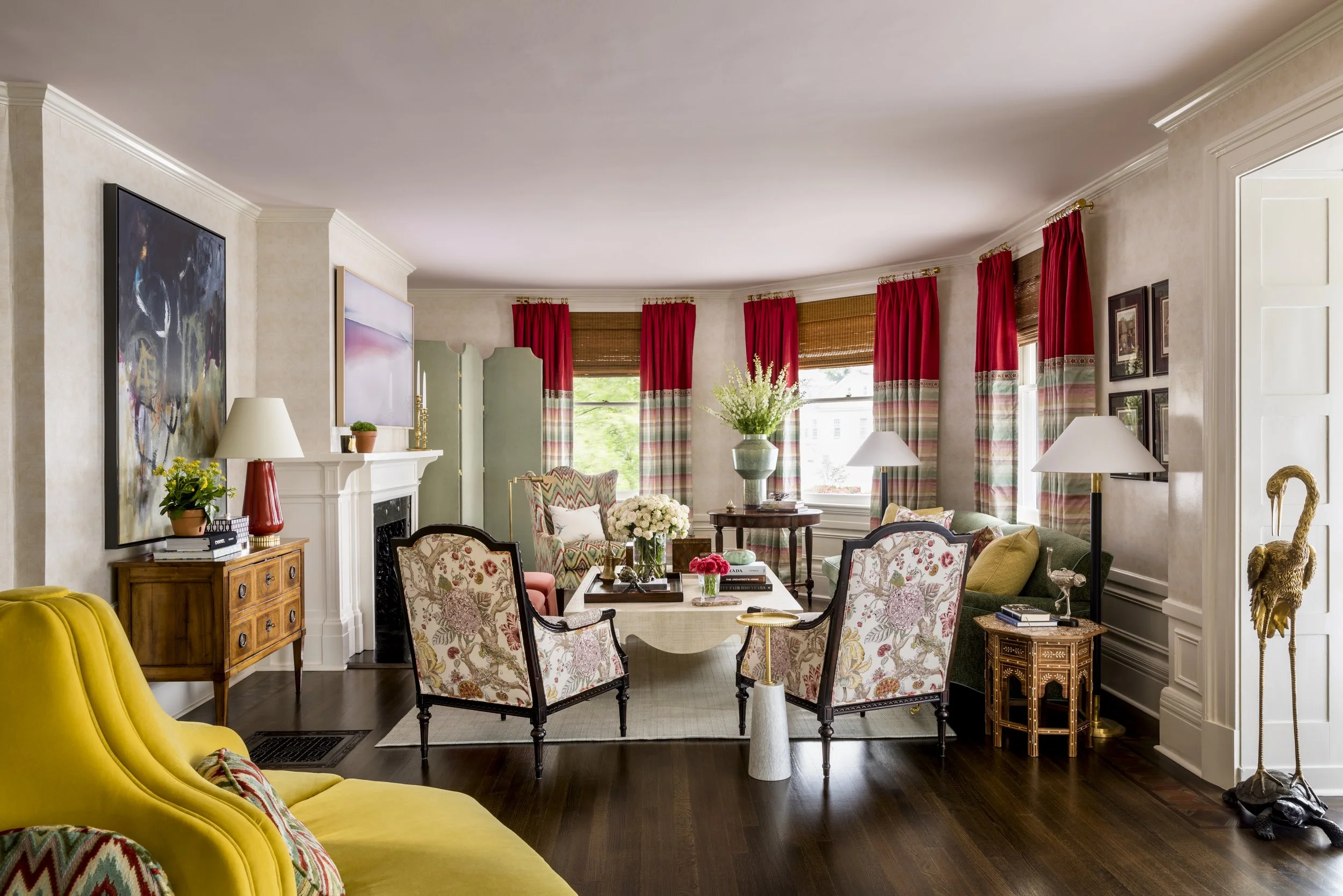 Living room with floral patterned chairs, a yellow sofa, paintings on the walls, red curtains, and a large window with greenery outside.
