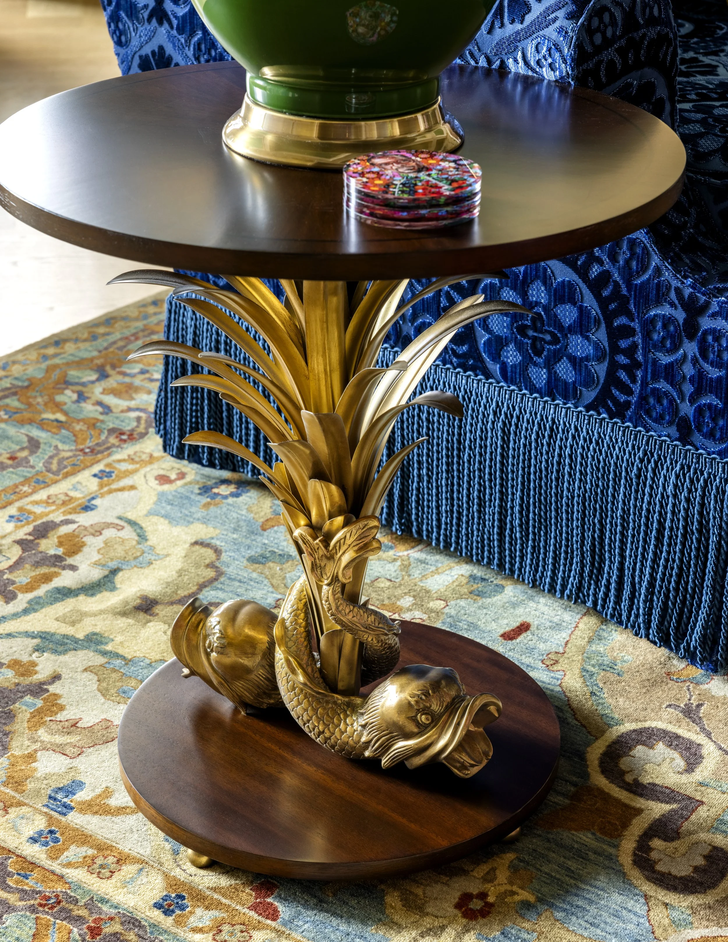 A decorative table with a gold fish sculpture base, a dark wooden surface, a green vase, and a stack of colorful coasters. A blue-patterned fabric and a patterned carpet are visible in the background.