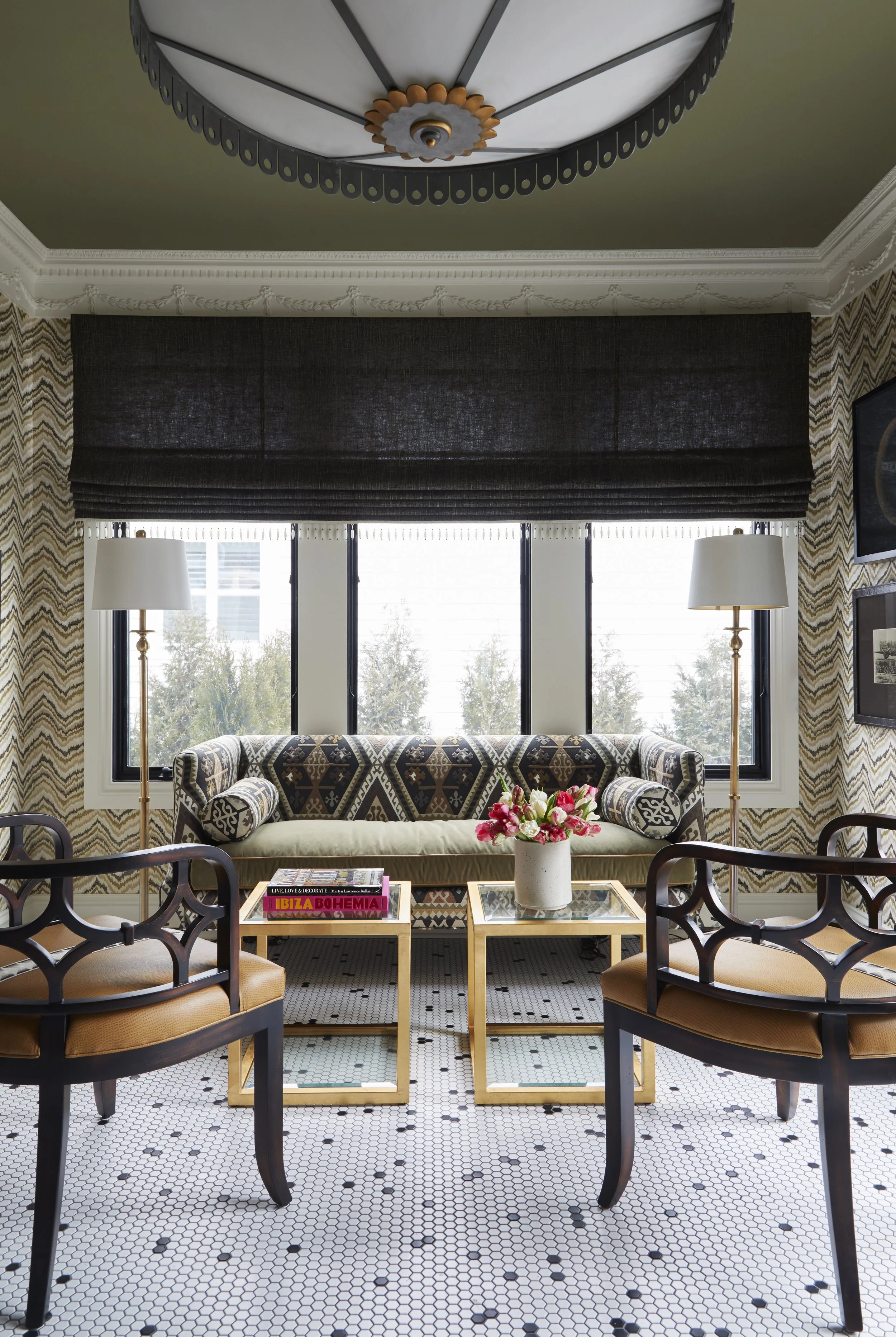 A living room with a brown sofa, two side tables with lamps, a coffee table with books and a flower vase, a large window with a black shade, and patterned wallpaper.