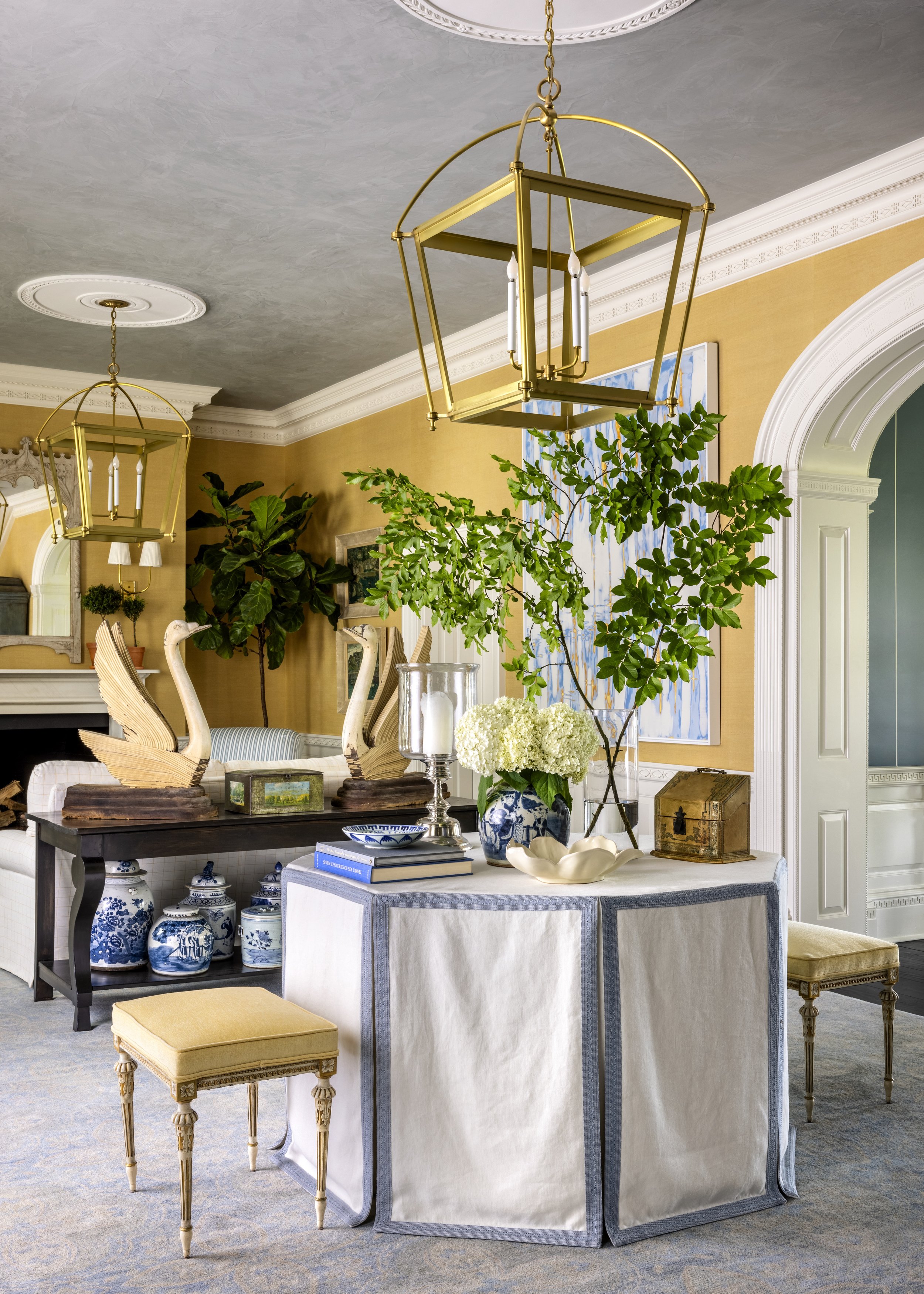 An interior dining room scene with a large round table, decorated with plants, vases, and decorative objects, with gold and blue accents, in front of a yellow wall and a large colorful painting. Two large gold chandeliers hang from the ceiling.