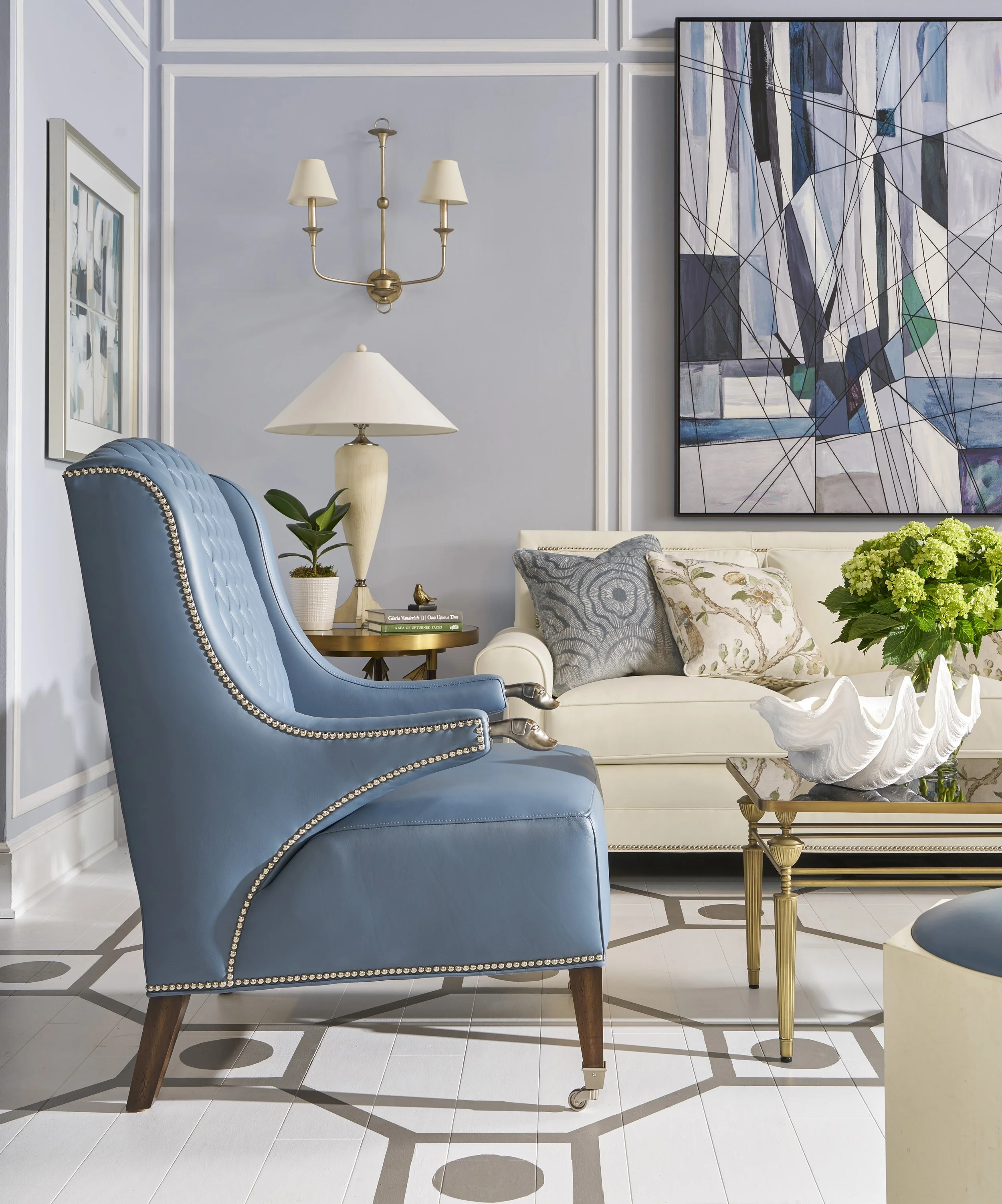 Interior of a living room with a blue upholstered chair with nailhead trim, a wooden side table with books and a beige lamp, floral pillows on a cream sofa, abstract art on the wall, a gold and white decorative bowl on the table, and a vibrant green 