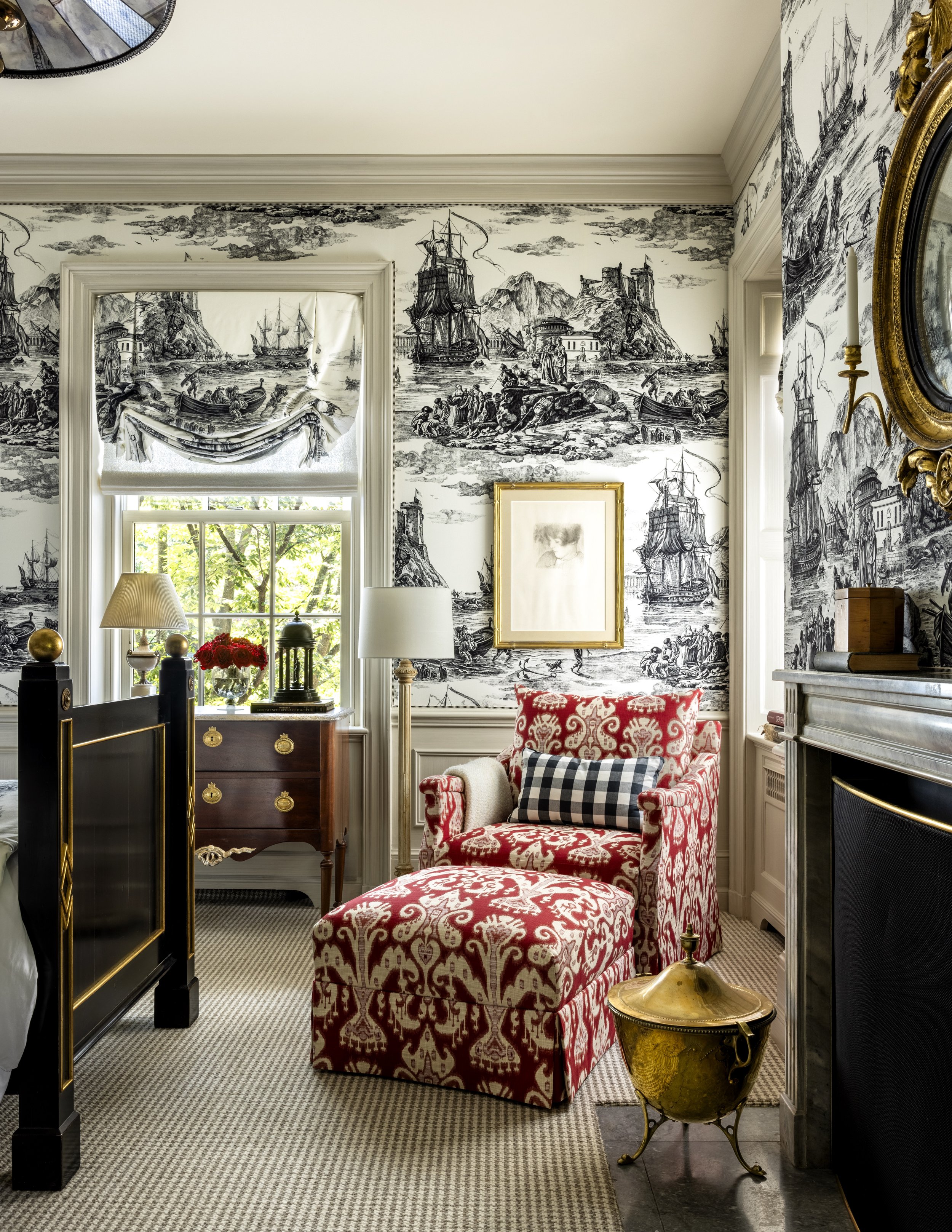 A cozy living room decorated in a classic style with a red and white patterned armchair, a matching footstool, a gold decorative vase, and black-and-white nautical-themed wallpaper depicting ships, the coast, and sea scenes. The room has large window