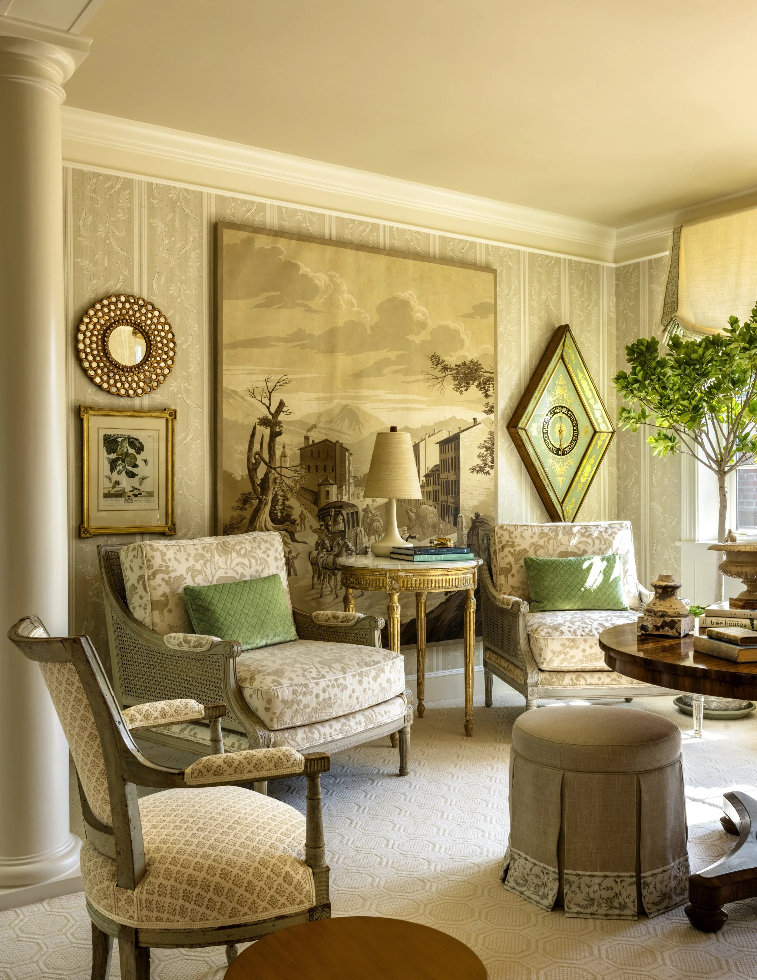 A cozy living room corner with beige and gold patterned furniture, including two armchairs with green pillows, a round coffee table, a beige ottoman, and decorative artwork on the walls, with natural light coming in from a window.