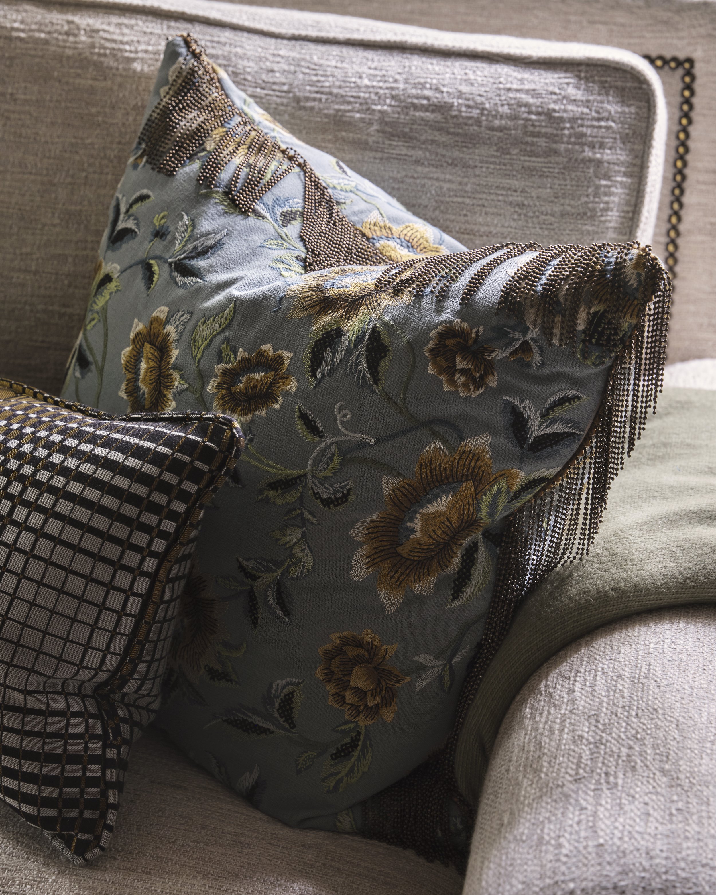 Close-up of a decorative pillow with floral embroidery and beaded fringe, resting against a upholstered chair with a textured beige fabric.