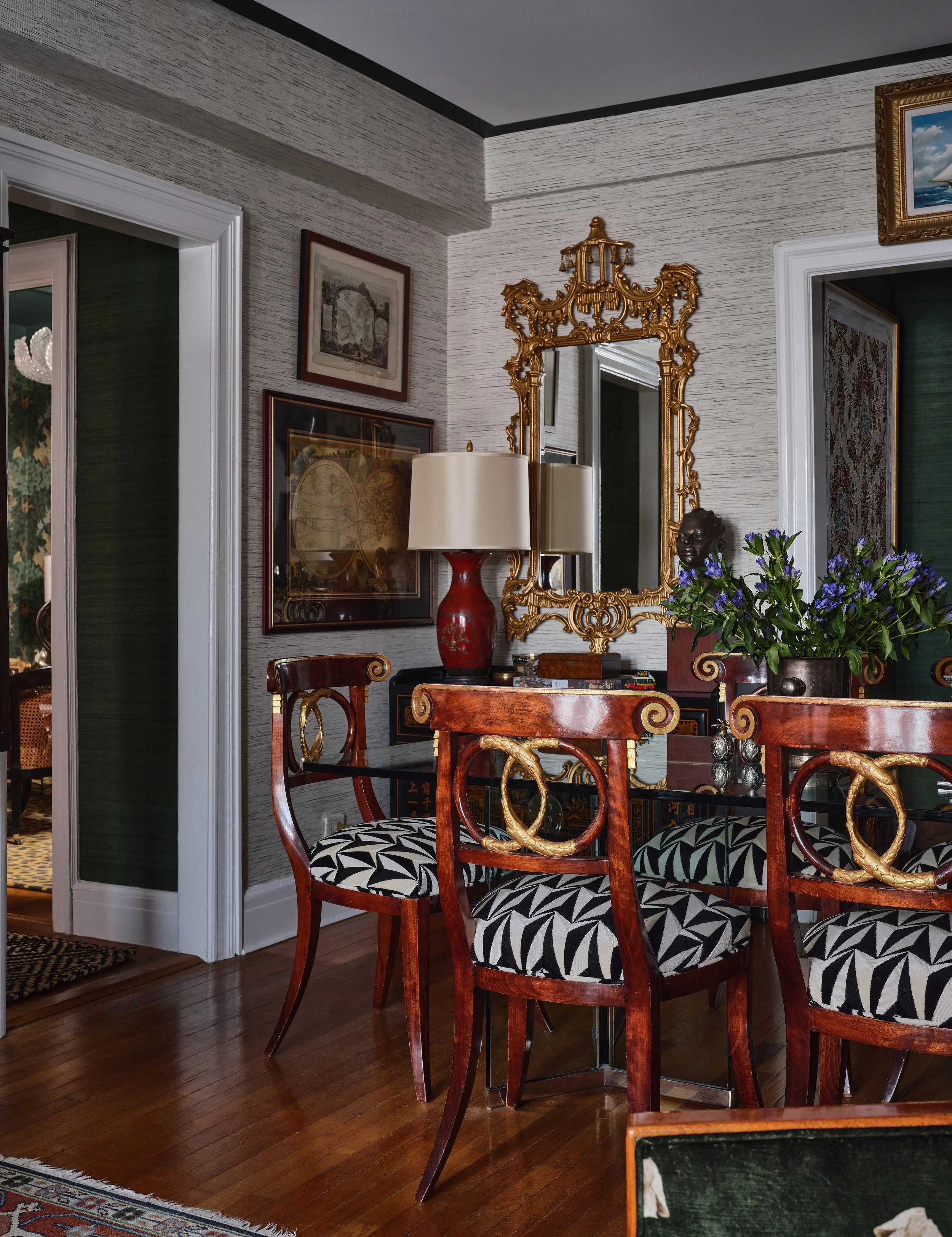 A room with wooden chairs featuring black and white geometric patterned cushions, a large ornate gold mirror, a red table lamp, framed artwork on the wall, a bouquet of purple flowers in a dark pot, and a dark wooden table.