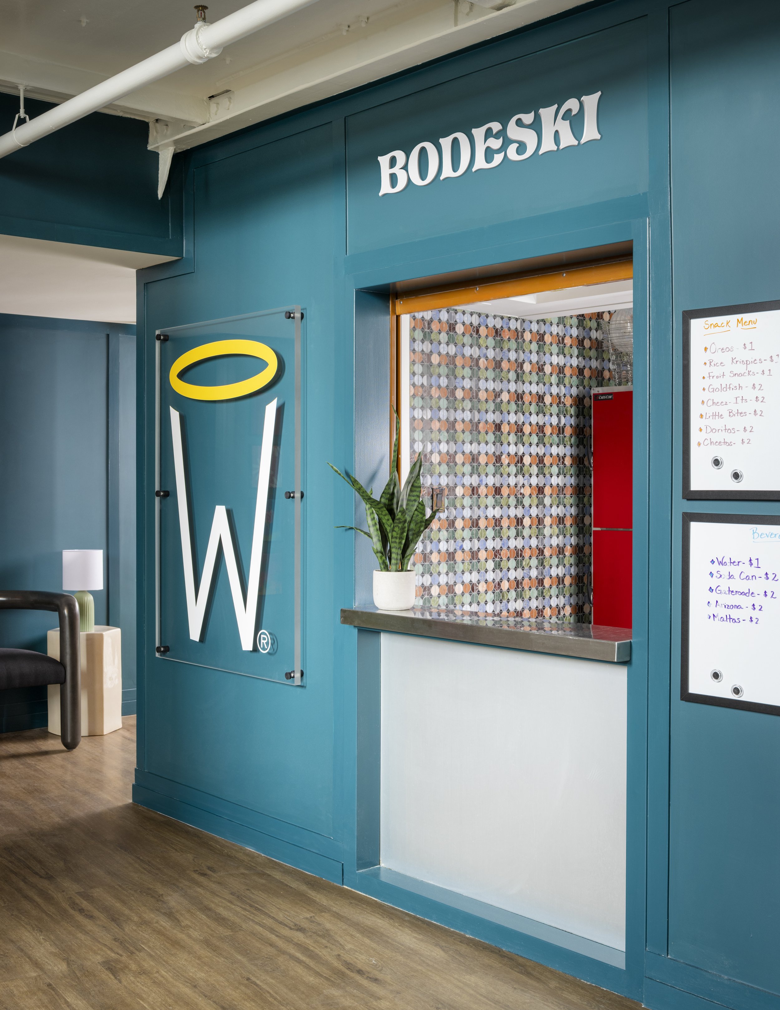 Interior of a modern building with a teal-colored wall and a sign that reads 'BODESKI'. A food counter with a patterned tile wall behind it, a potted plant on the counter, and a white menu board with handwritten items and prices. Part of a small seat