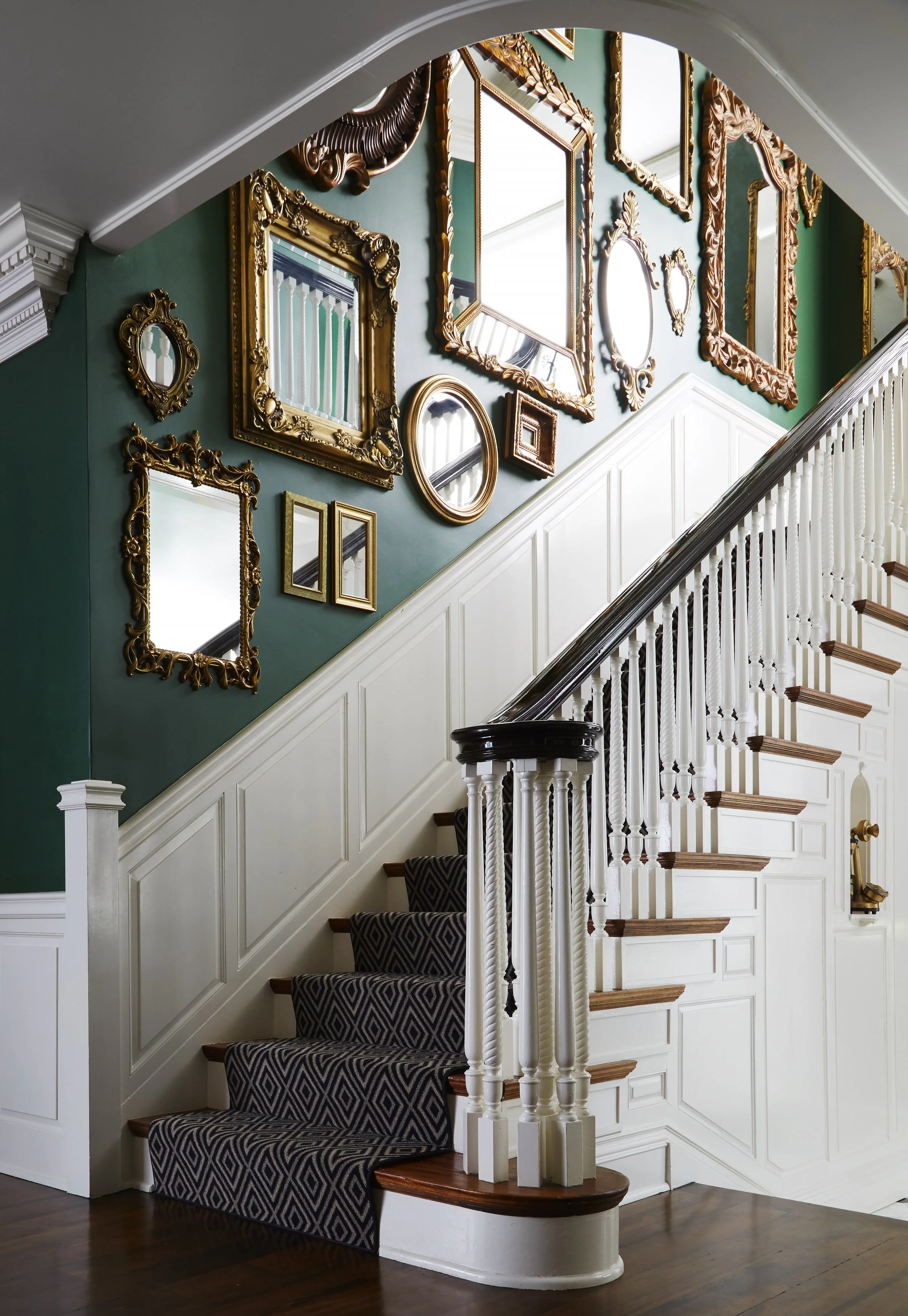 Interior view of a staircase with decorative mirrors on a green wall, featuring gold and wooden frames, and a staircase with a dark handrail and patterned carpet.