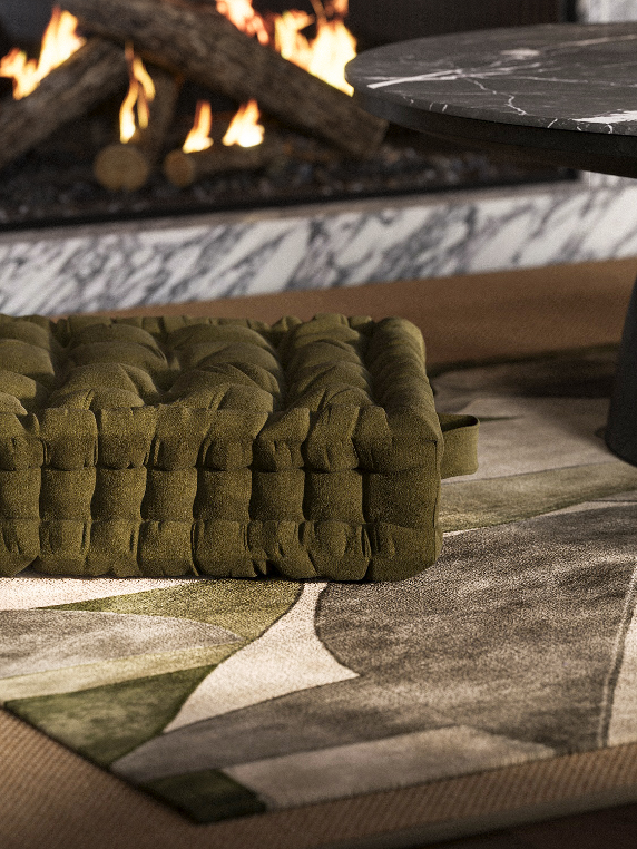 Close-up of a green textured cushion on a patterned carpet near a fireplace with flames and logs.
