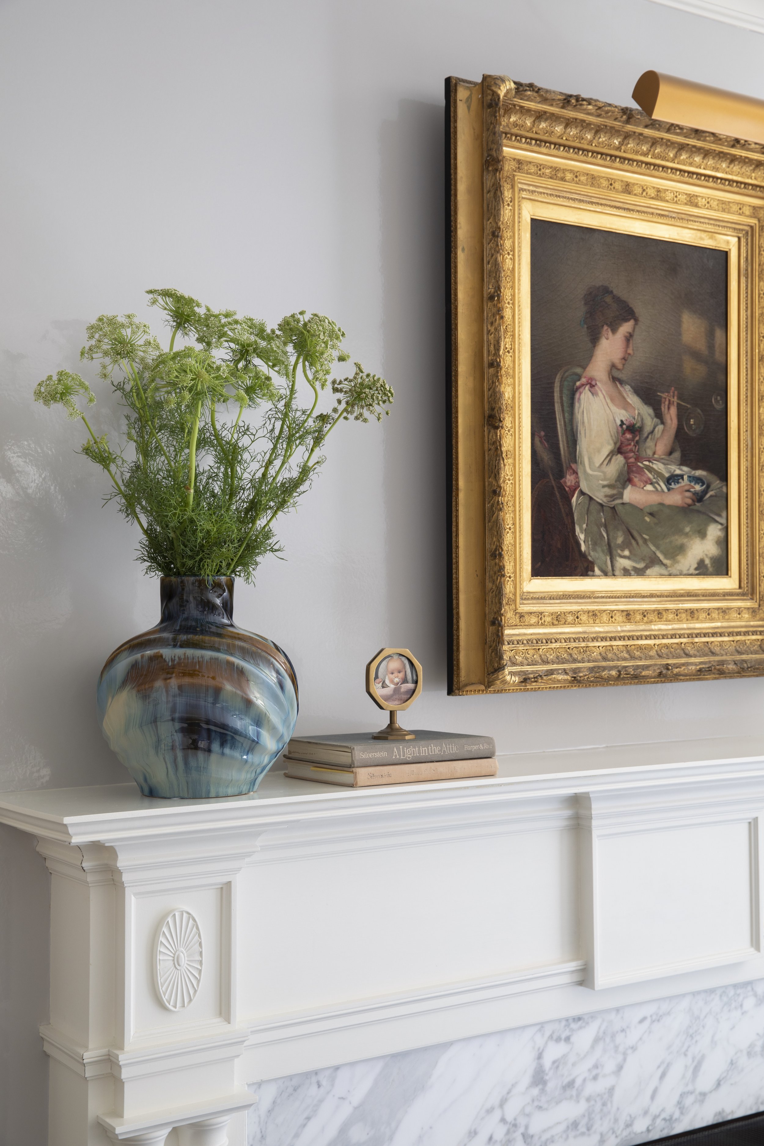 Decorative white fireplace mantel with a large blue and brown ceramic vase containing green wildflowers on the left, a stack of books topped with a small photo frame on the right, and a framed painting of a woman playing chess hanging on the wall abo
