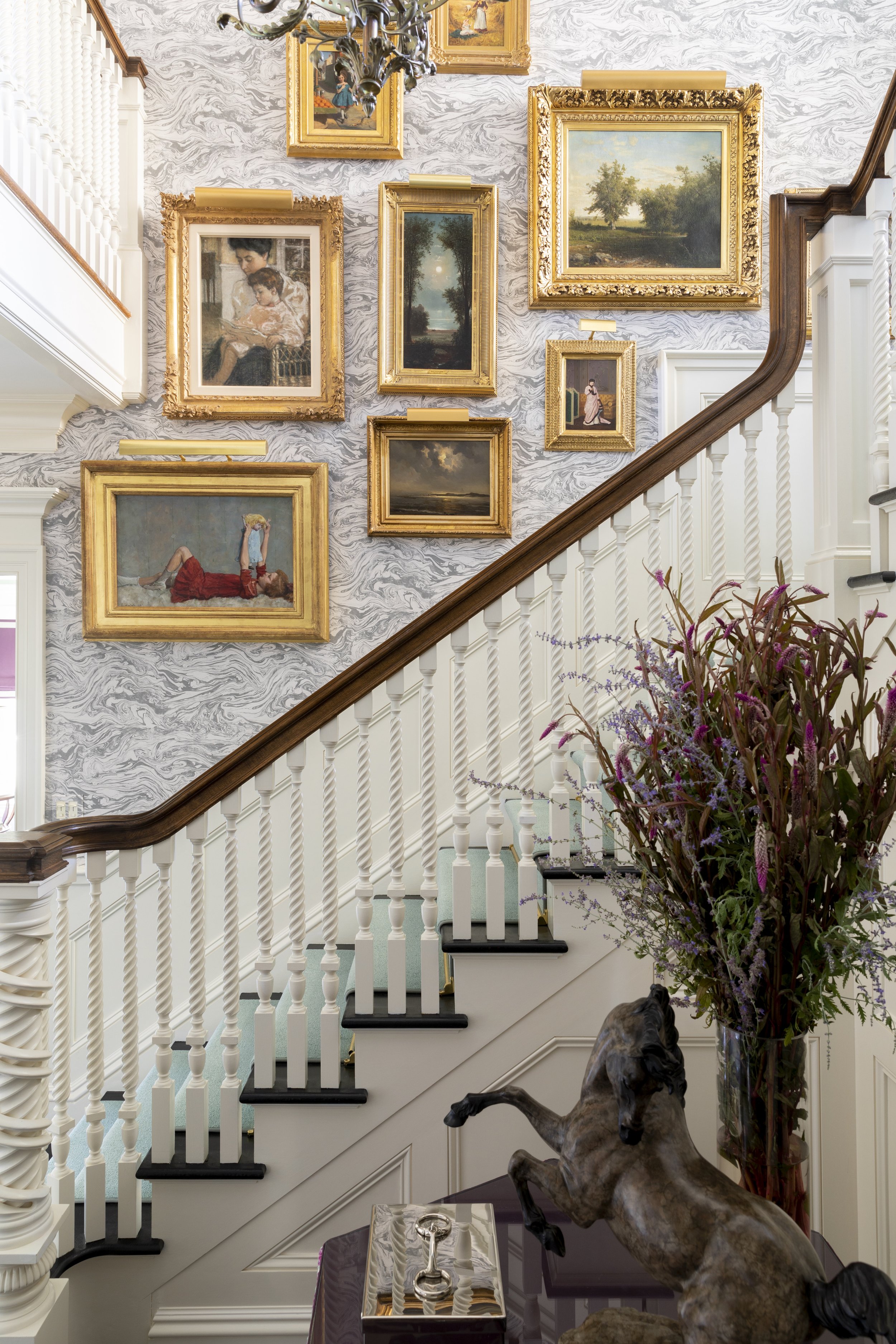 An elegant staircase with white balusters and a dark wooden handrail, decorated with framed paintings in ornate gold frames on a marbled-patterned wall. A bronze horse sculpture and a large vase with flowers are near the staircase. A small table hold