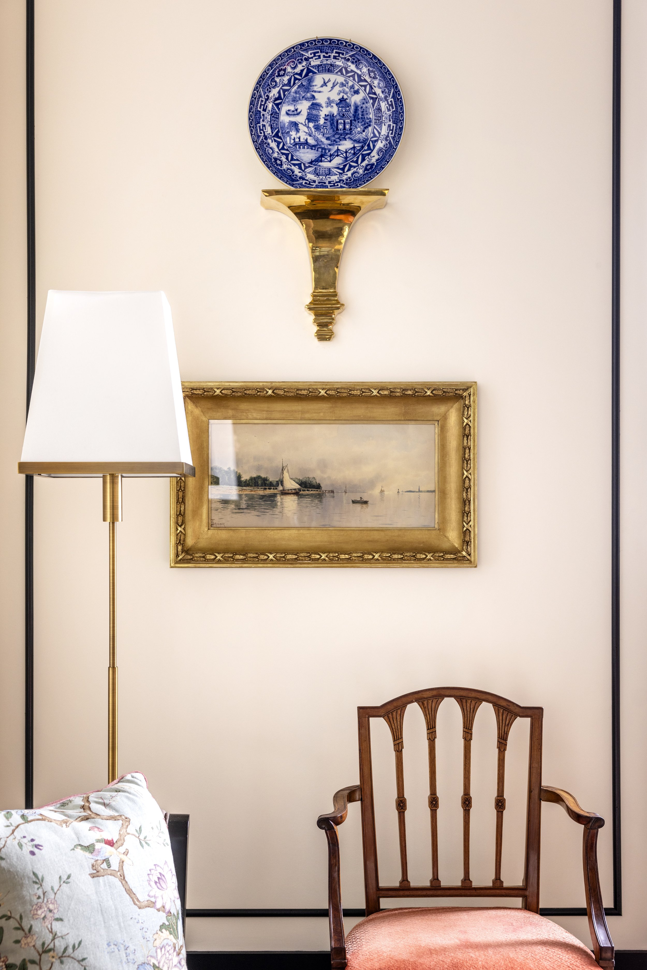 Interior view of a room with a wooden chair, a white lamp, a framed painting of a sailboat on water, a decorative wall piece with a blue and white design, and a floral pillow.