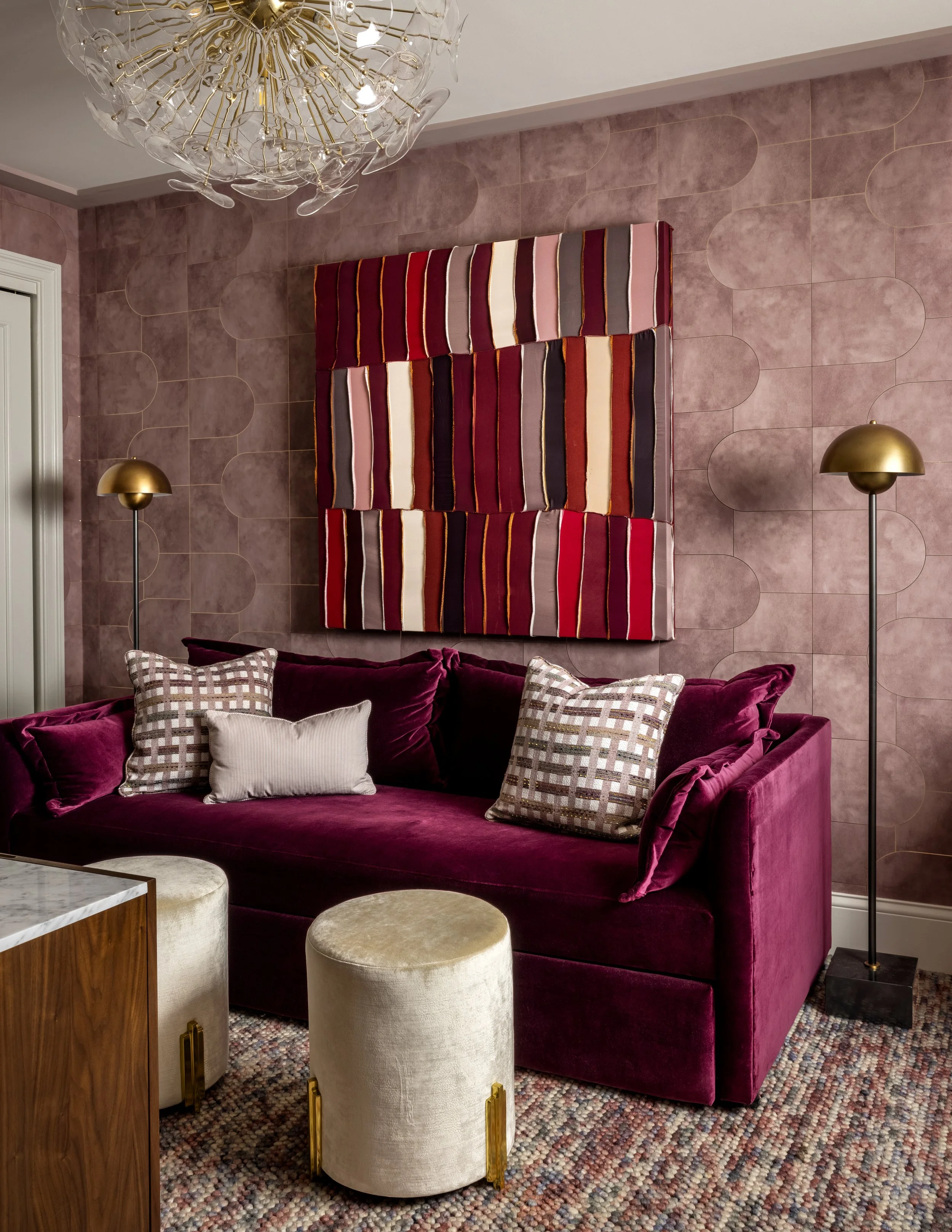 A living room with a maroon velvet sofa adorned with checkered and solid throw pillows. Behind the sofa, a textured pink wall features a large, vertical, striped textile art piece with shades of red, pink, beige, and gray. Two floor lamps with gold d