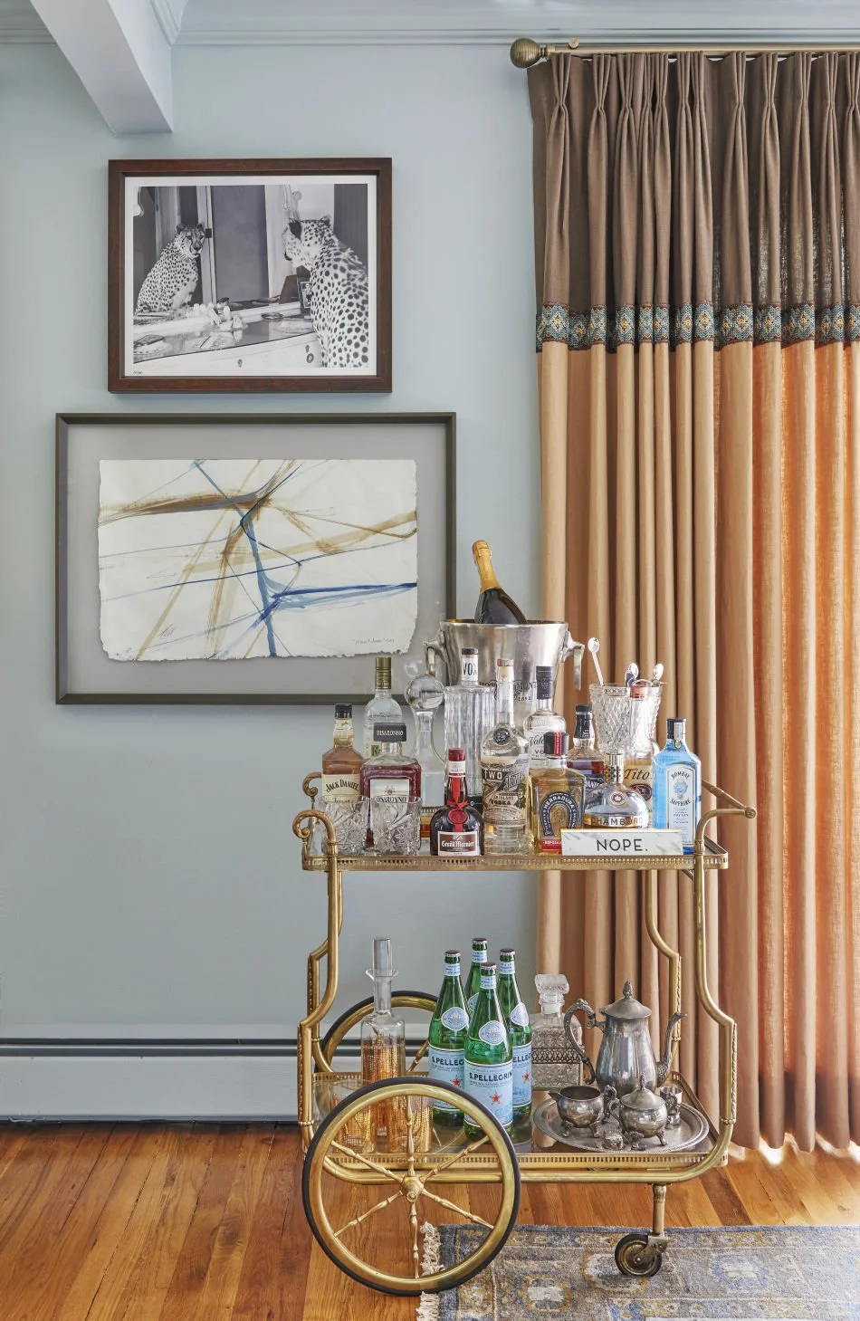A gold bar cart with bottles of alcohol, mixers, and glassware, positioned next to beige and brown curtains. Two framed pieces of art are hung on the light blue wall behind the cart, and a black and white photo of two leopards is on top. The cart has