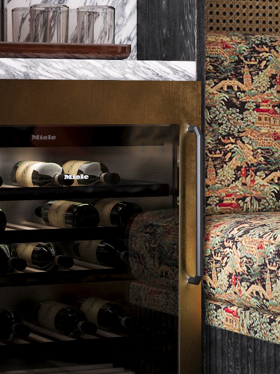 Wine bottles on shelves inside a glass wine cooler next to a patterned upholstered chair.