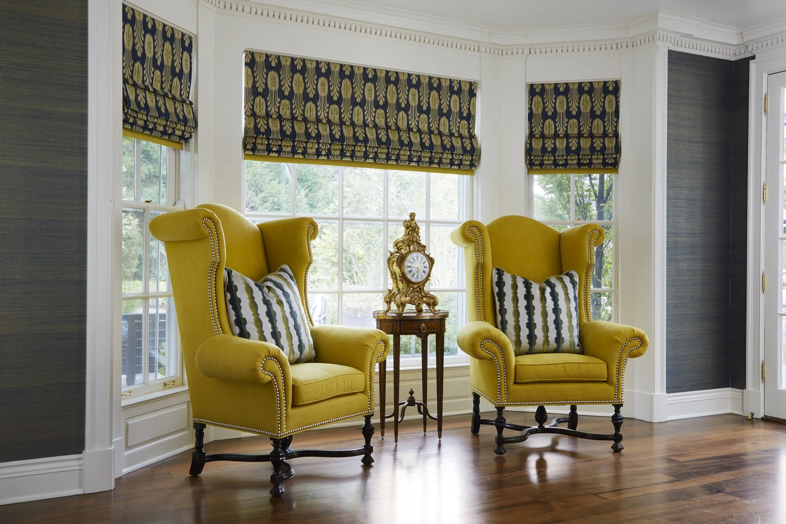 Living room with two yellow armchairs, a small wooden table with a clock, windows with patterned blue and gold roman shades, and hardwood floors.