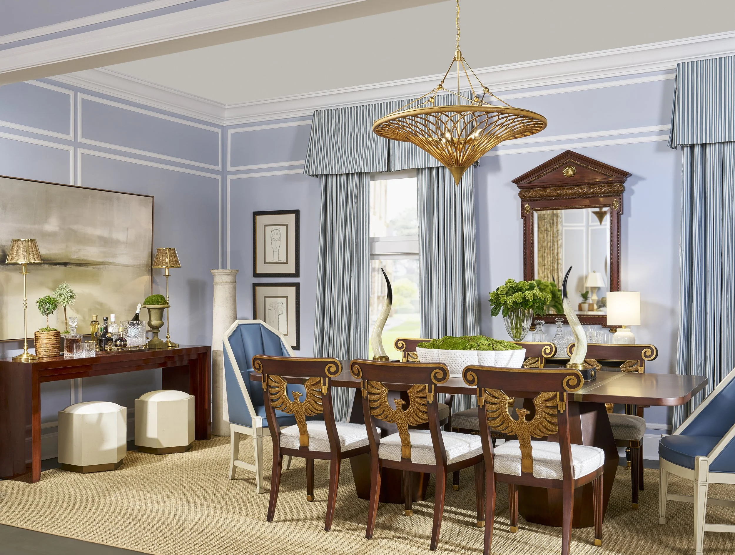 A formal dining room with a large wooden dining table surrounded by ornate wooden chairs. The room features light blue walls with white decorative trim, striped curtains, and a gold contemporary chandelier hanging from the ceiling. There are various 