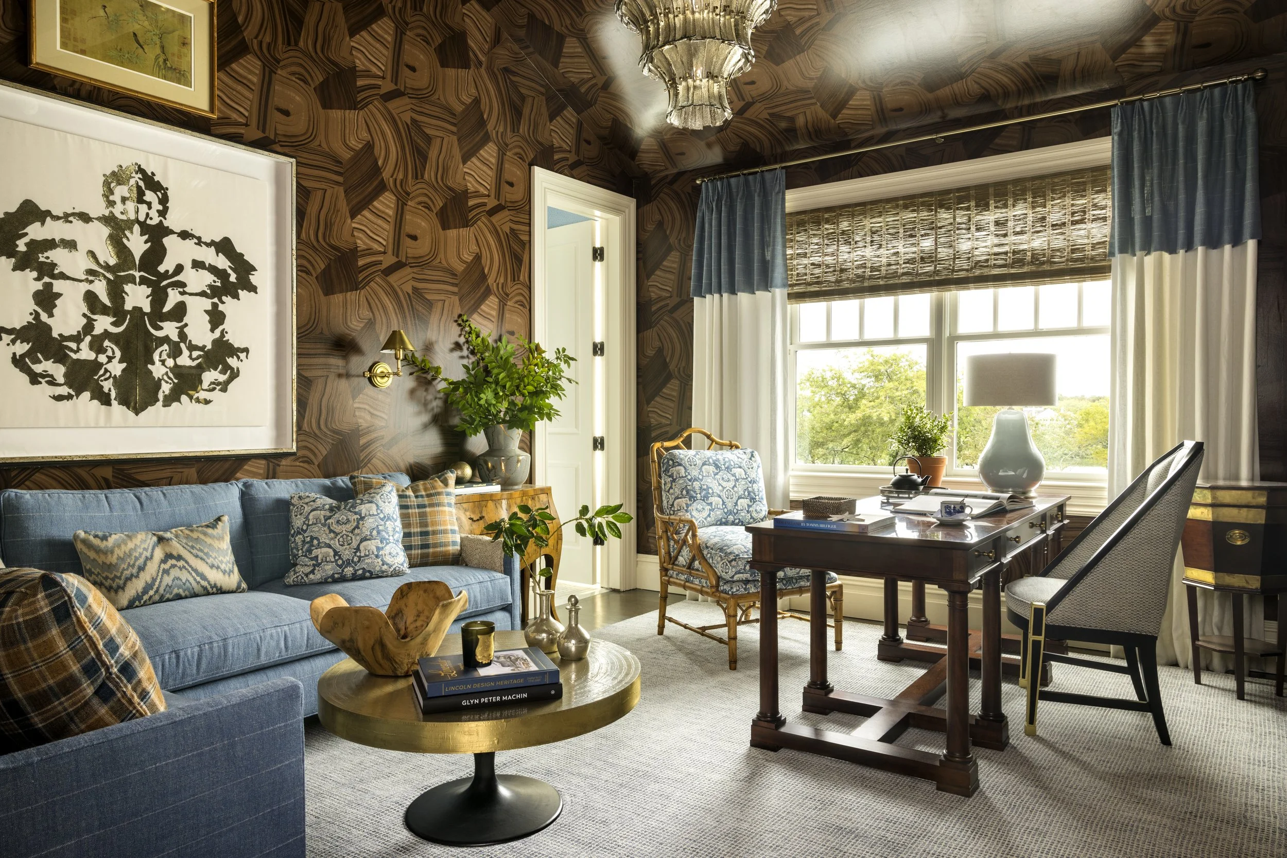 Living room with wooden patterned wallpaper, large window with blue and white curtains, blue sofa with patterned pillows, wooden coffee table with books and decorative items, armchair with blue and white fabric, potted plants, art on the wall, chande