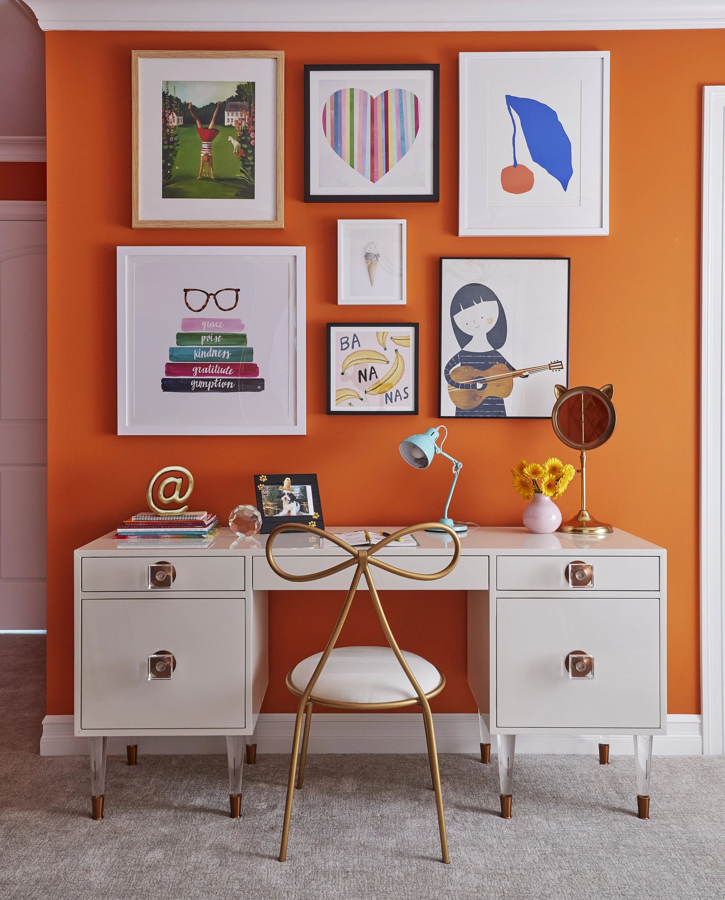 A white desk with gold and white legs, topped with various items including a yellow desk lamp, a pink vase with yellow flowers, a photo frame, a small black book, a notebook, and a glass paperweight. There is a gold chair with a rounded back and whit