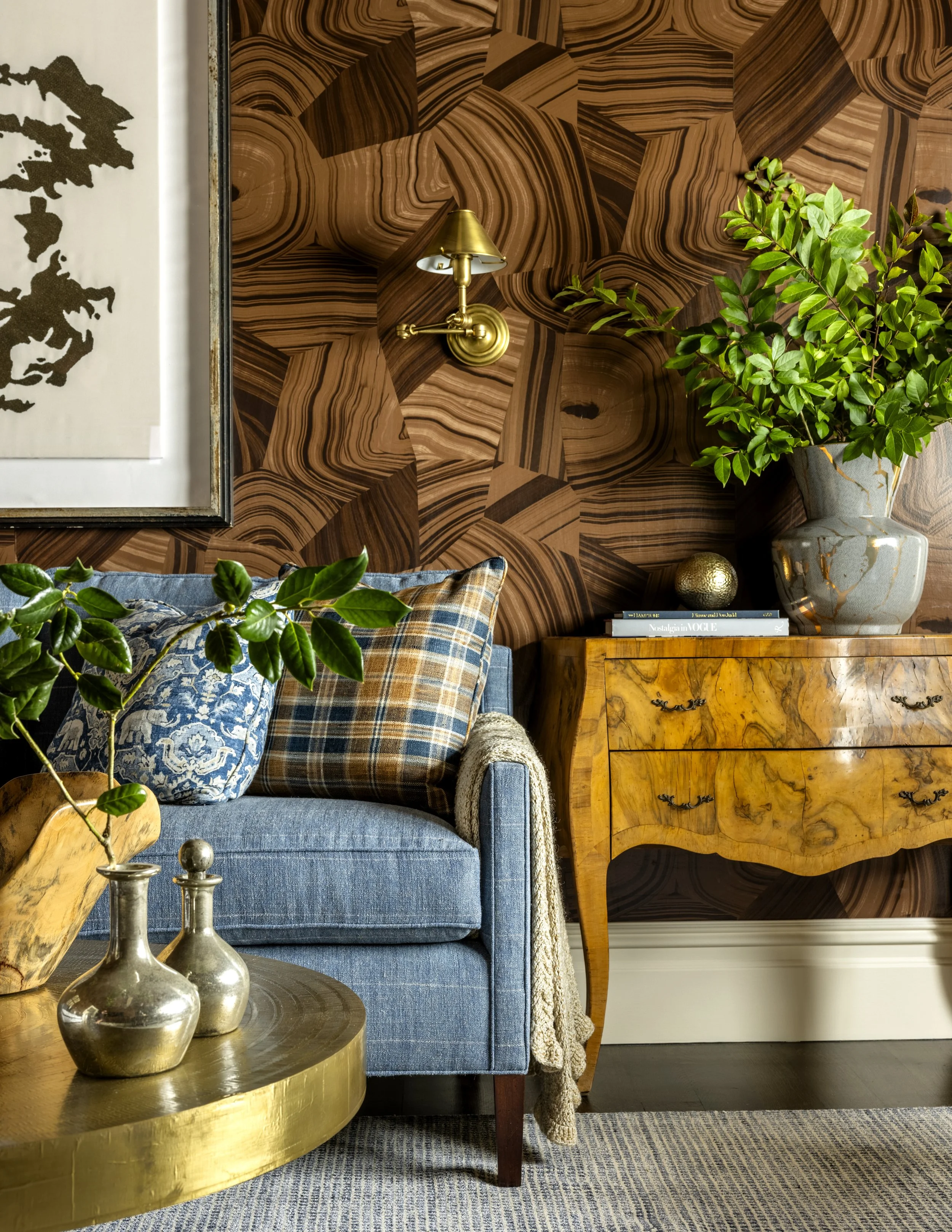Living room with a blue sofa, plaid pillows, a wooden coffee table with metal vases and a decorative object, a green plant in a patterned vase, a gold wall lamp, intricate wood wall paneling, and a painting on the wall.